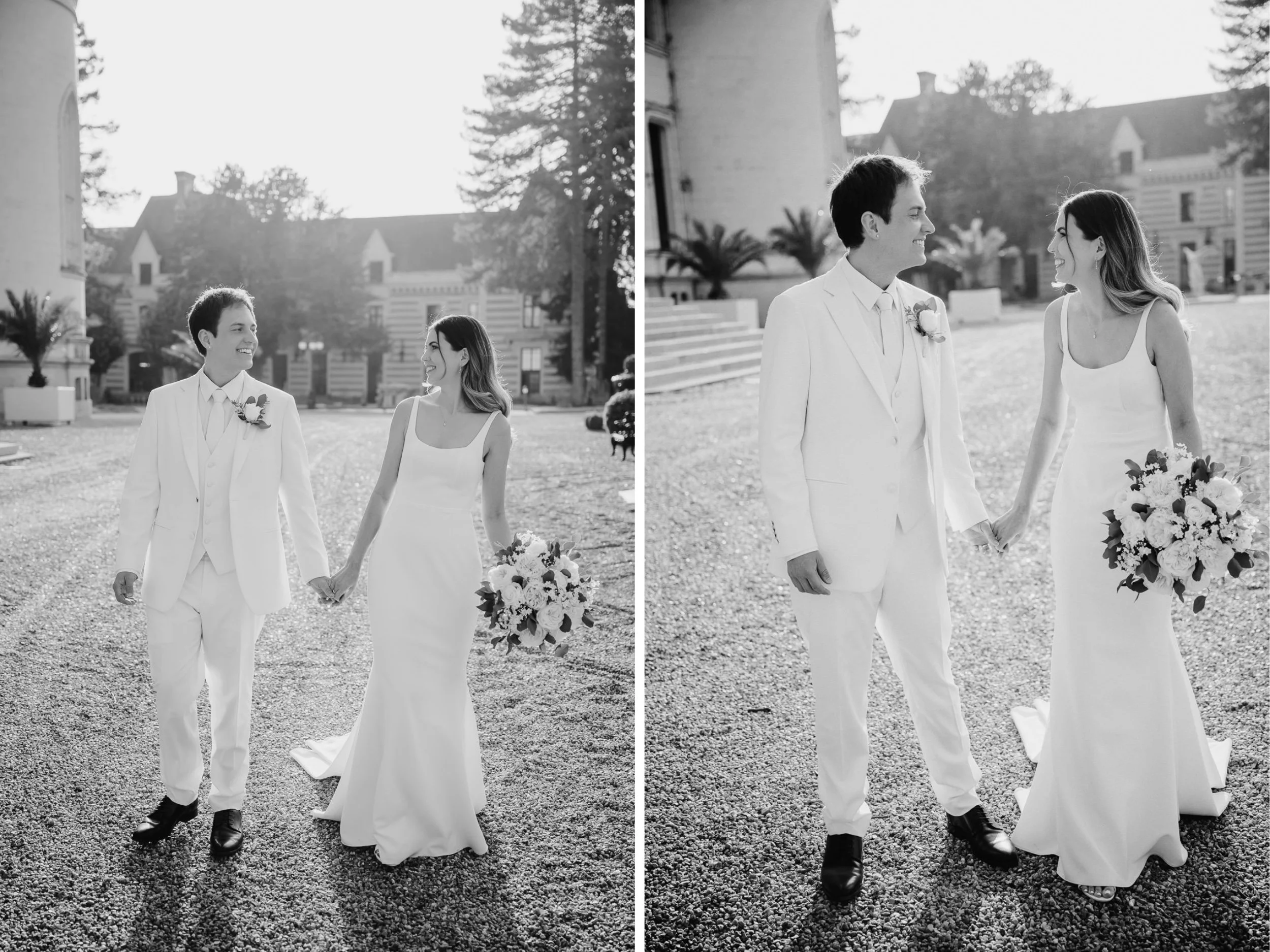 Two women in wedding dresses walking outside holding hands, smiling at each other, with one woman holding a bouquet of flowers, and a man in a white suit walking alongside them, in front of a large building with trees in the background.
