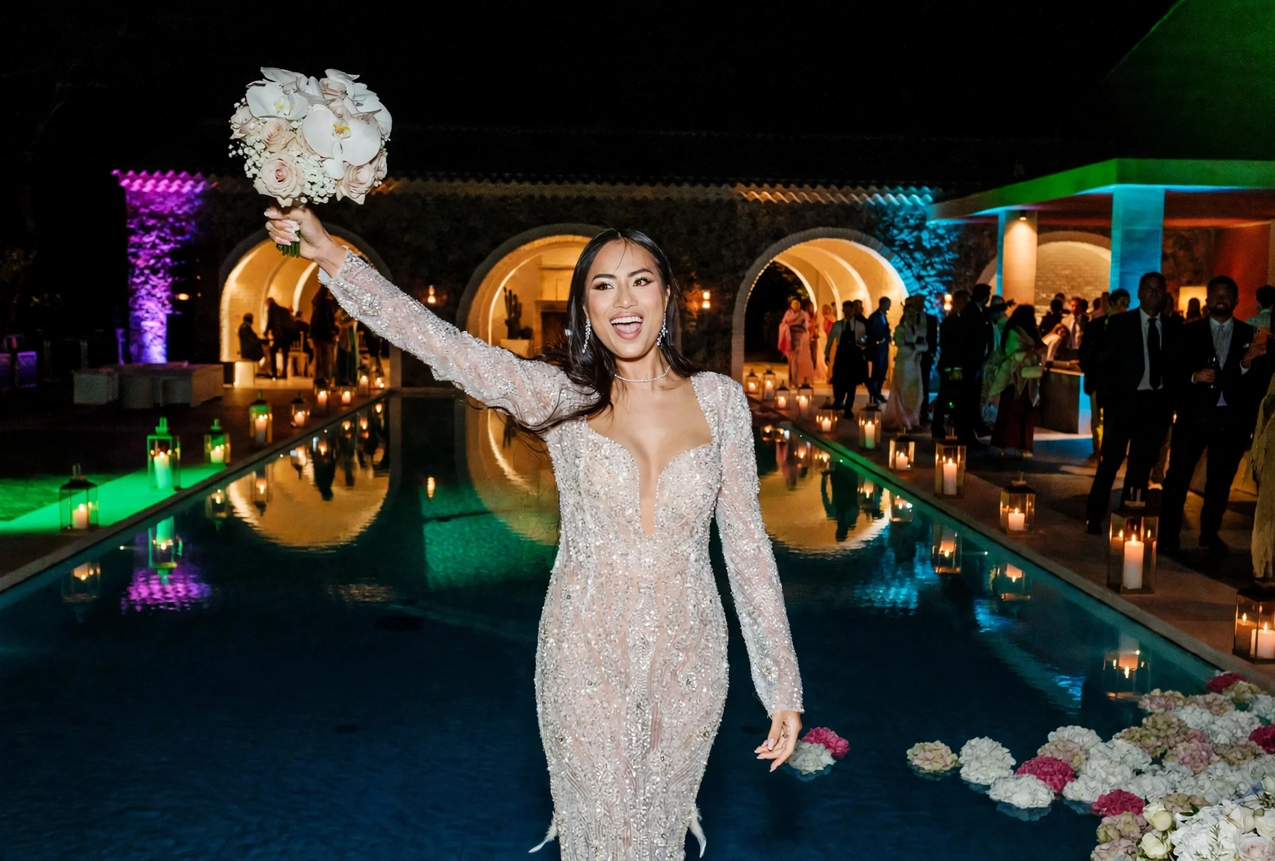 Joyful bride in sparkling sequin gown with orchid bouquet by candlelit pool at luxury night wedding
