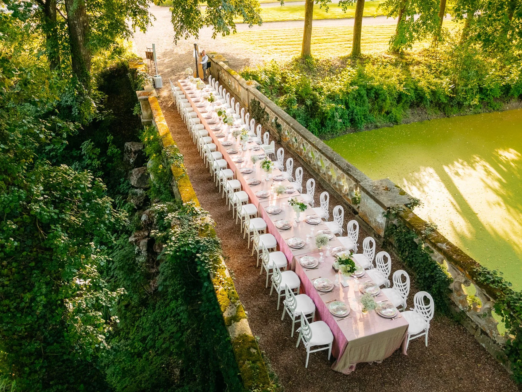 Long outdoor dining table with pink tablecloth set for a meal, decorated with flowers, situated on a pathway along a pond with green algae, surrounded by tall trees and lush greenery.