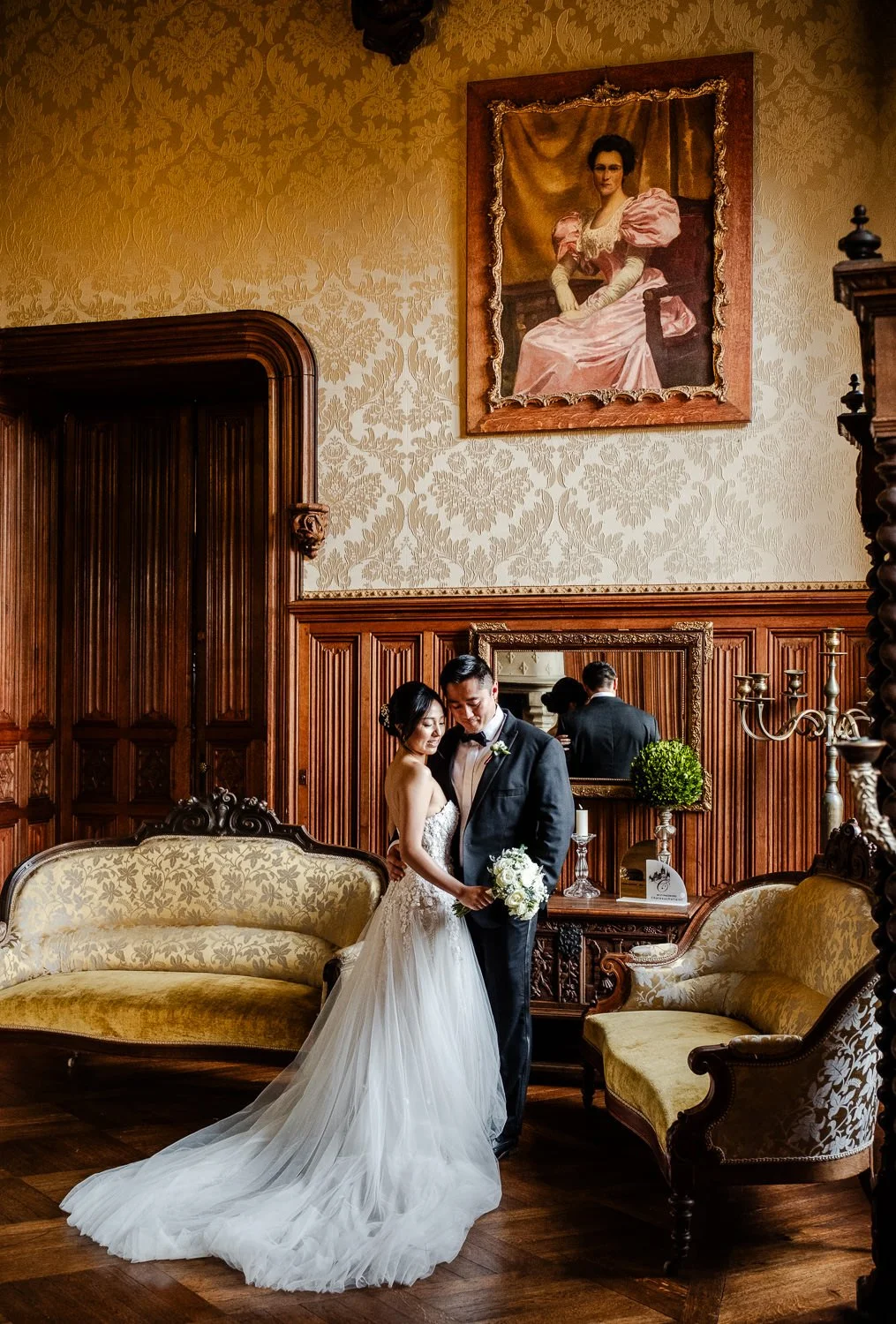 A bride and groom in wedding attire standing close together in an elegant, vintage-style room with ornate wood paneling, a large portrait of a woman in a pink dress hanging on the wall, and a mirror reflecting part of their embrace.