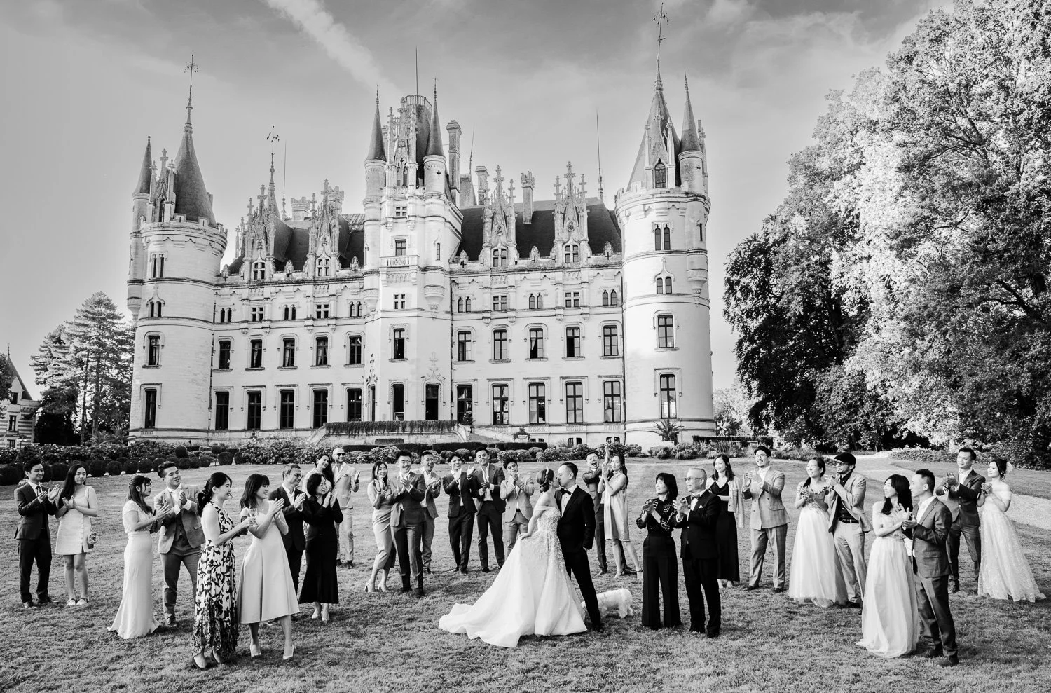 A wedding party taking photos in front of a large castle with spires, surrounded by trees.