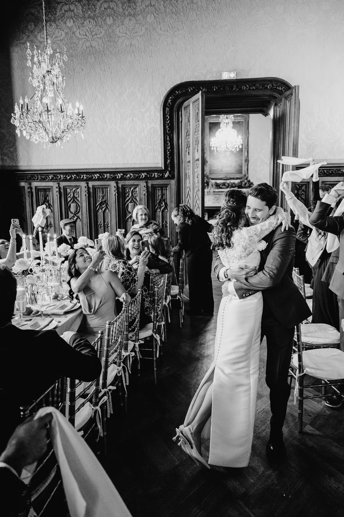 Black and white photo of a wedding reception with a bride and groom hugging in front of a long dining table filled with guests, elegant chandeliers hang from the ceiling, and a chandelier is reflected in a mirror in the background.