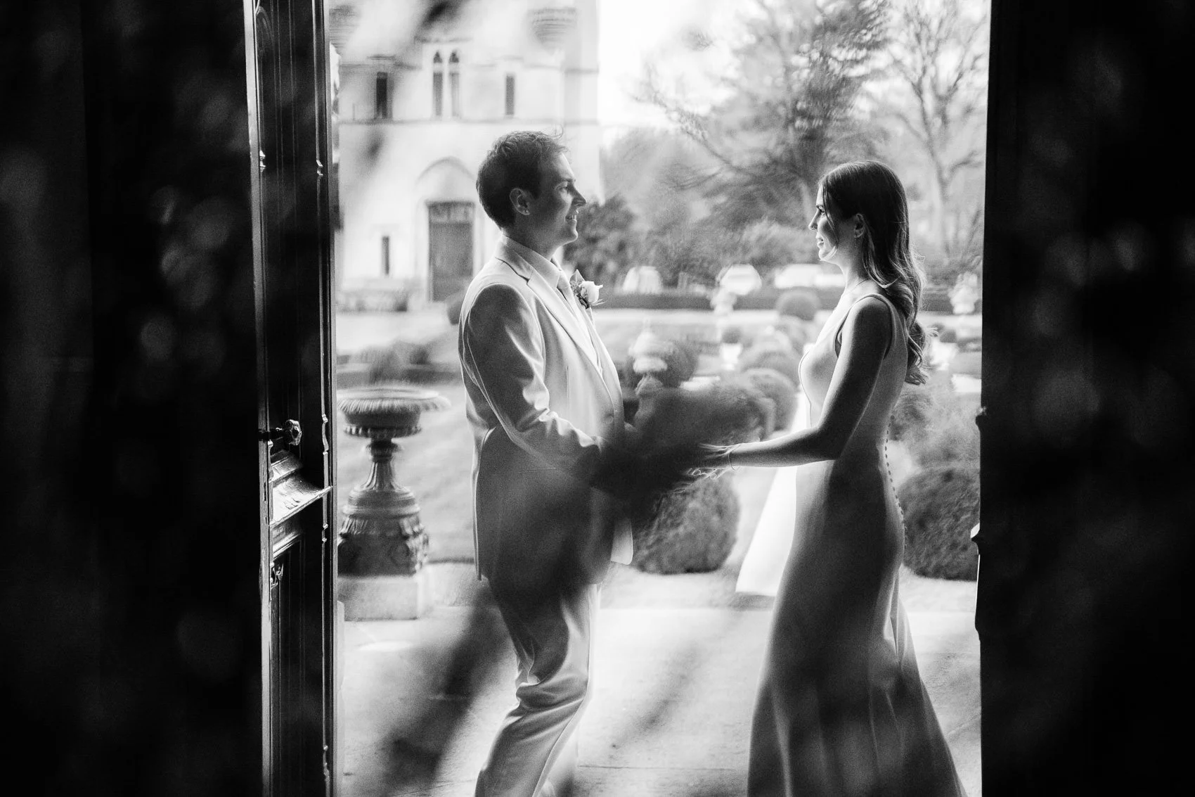 A black and white photo of a bride and groom holding hands and gazing at each other in front of a glass door, with a garden and building visible outside.