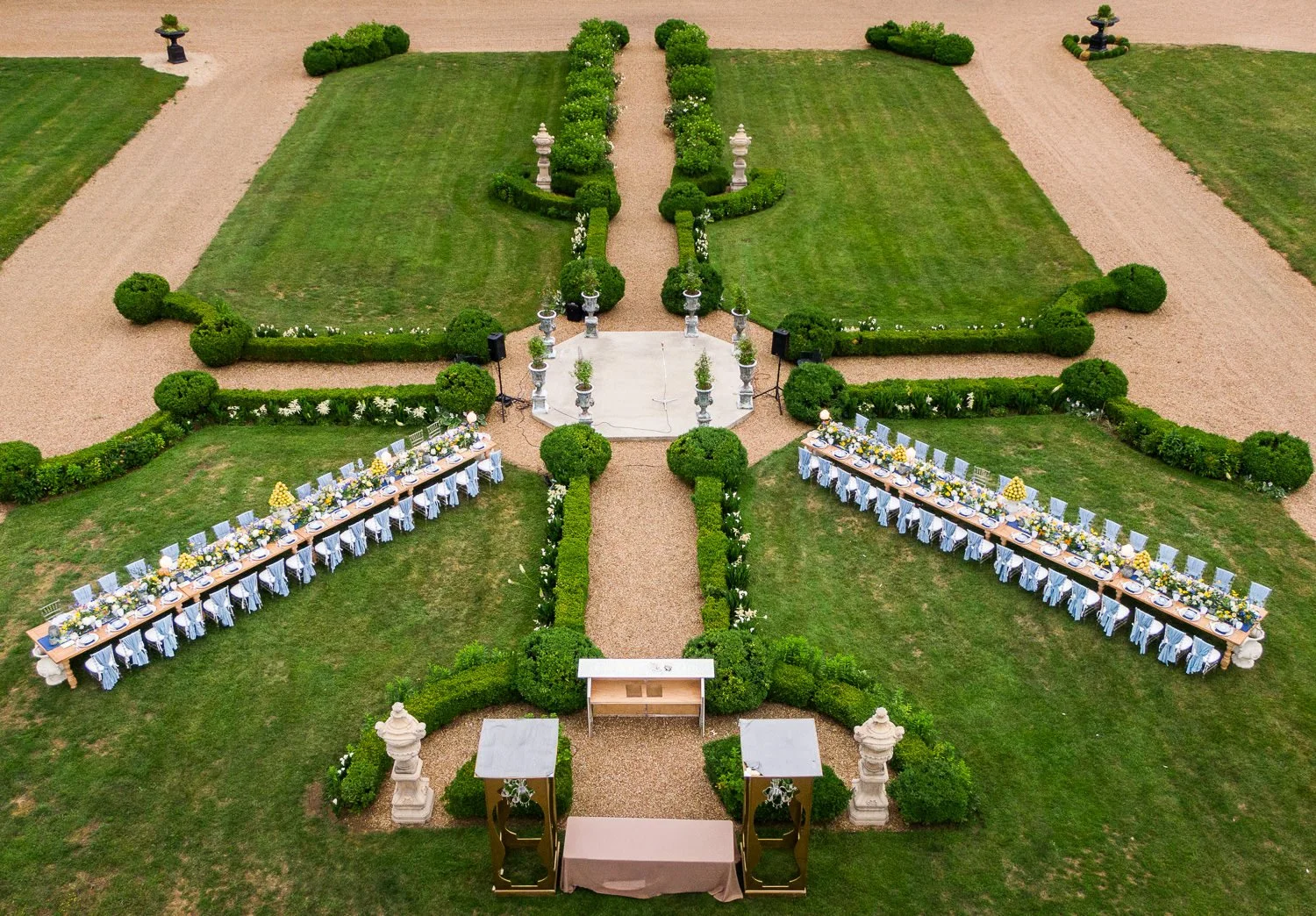 A garden setup for a wedding or event with long decorated tables, rounded hedges, and a pathway leading to a stage area with angel statues, surrounded by hedges and greenery.