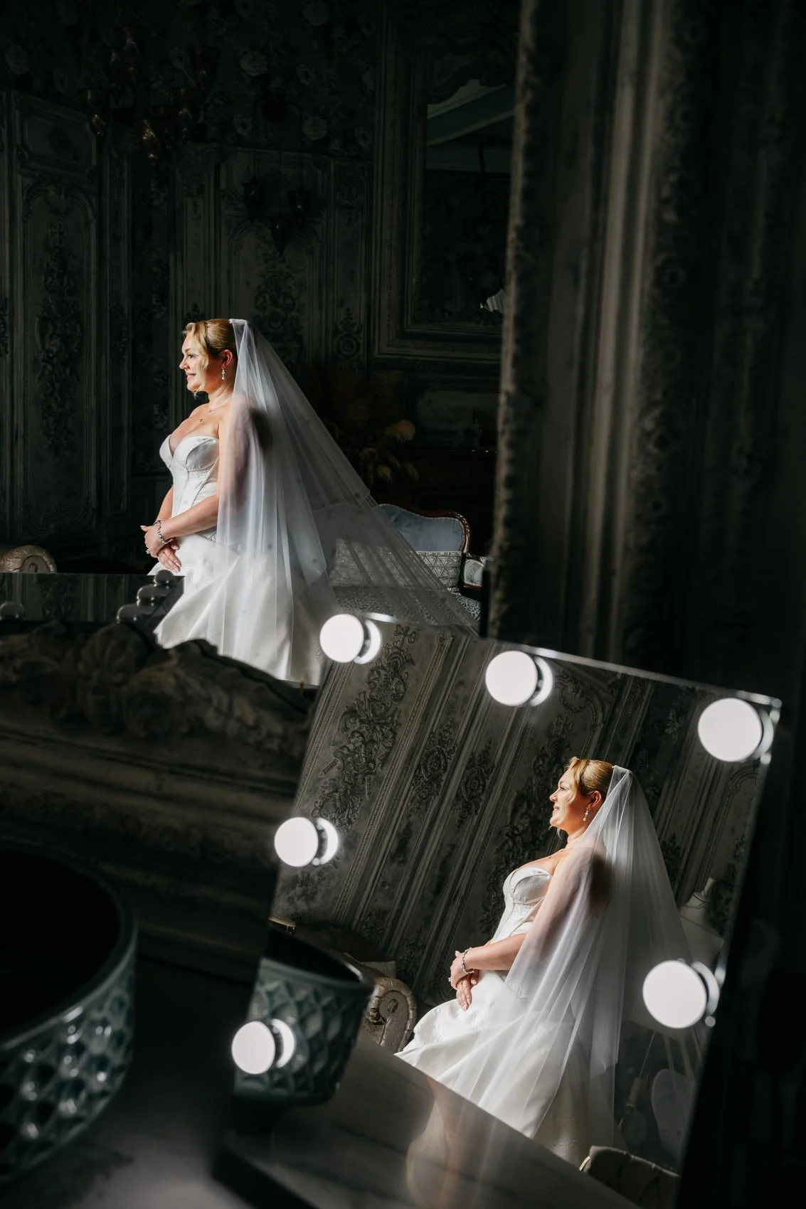 A bride in a white wedding dress and veil sitting in front of a mirror with lights. The mirror reflects her profile and ornate room details.