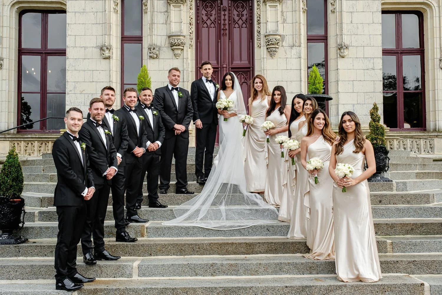 Group of bridesmaids and groomsmen on stairs in front of a historic building, attending a wedding. The bride and groom are in the center, with the bride wearing a white wedding gown and veil, holding a bouquet, and the groom in a black tuxedo. Brides