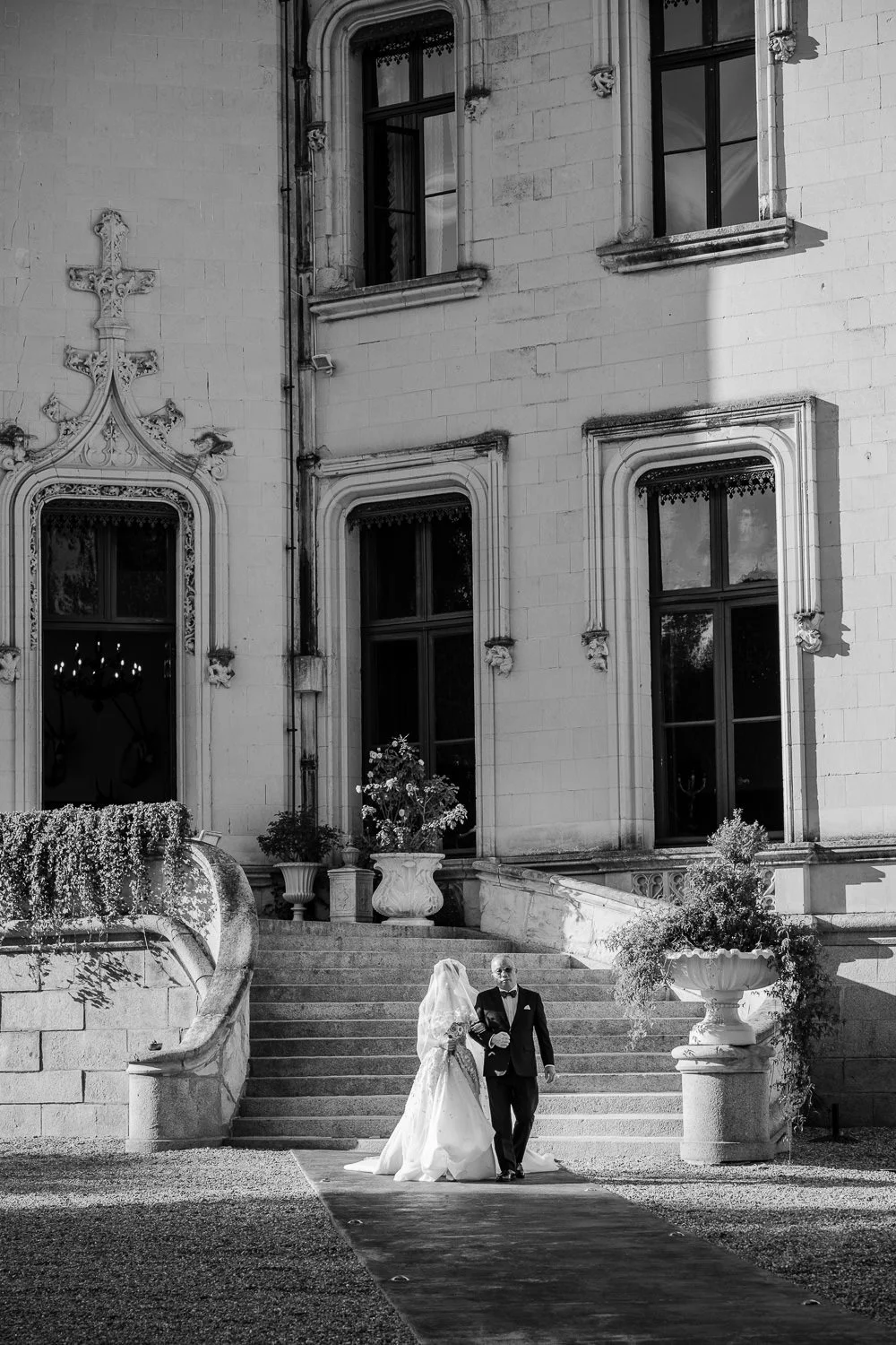 A bride and groom walking down an outdoor aisle in front of a historic stone building with gothic windows and ornate details, during their wedding ceremony.