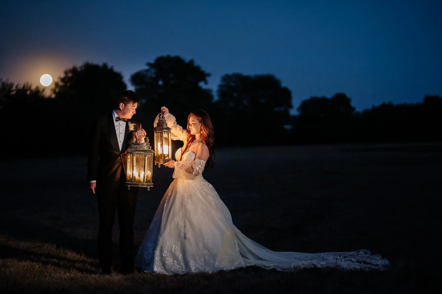 A wedding couple at night holding lanterns, with the moon and trees in the background.