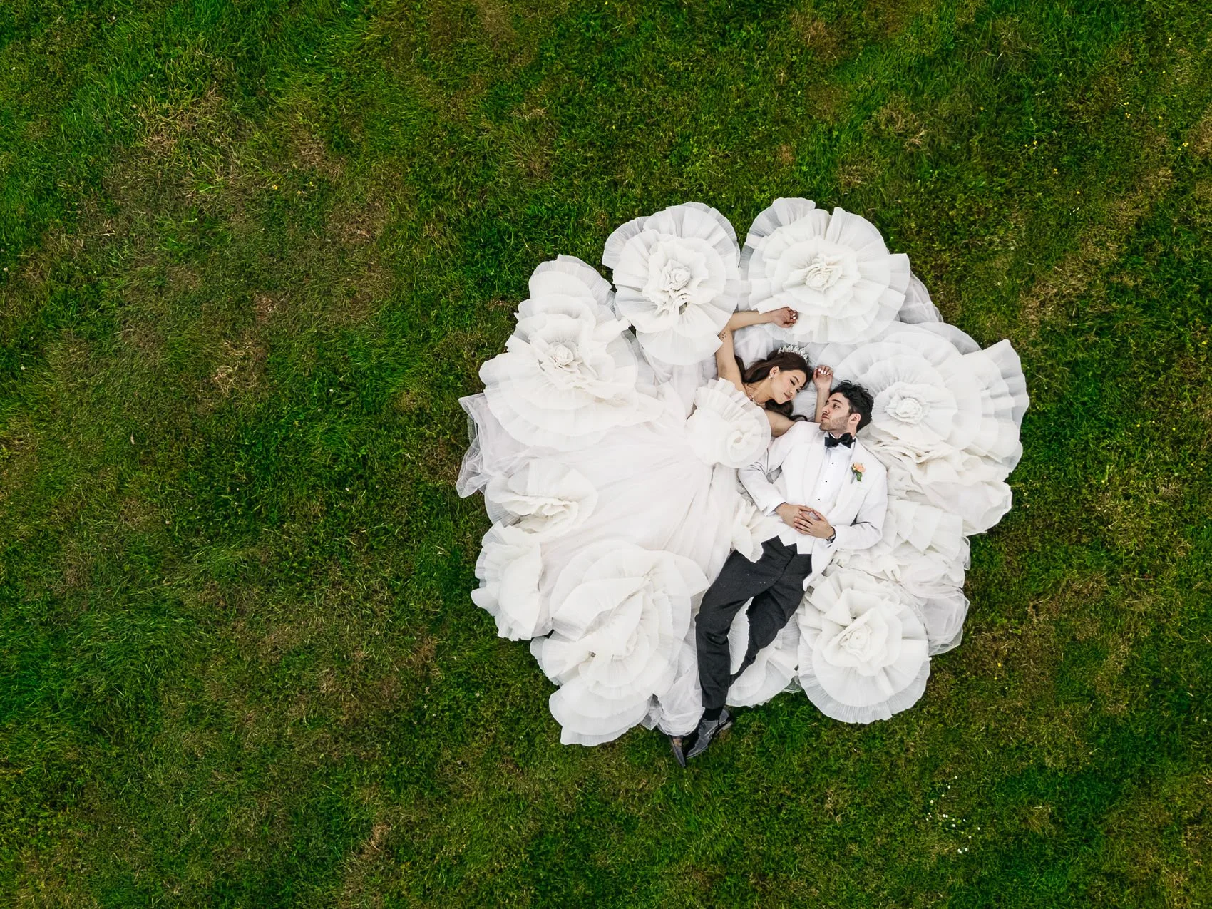 A bride and groom lying on green grass surrounded by large white fabric flowers, seen from above, at their wedding.