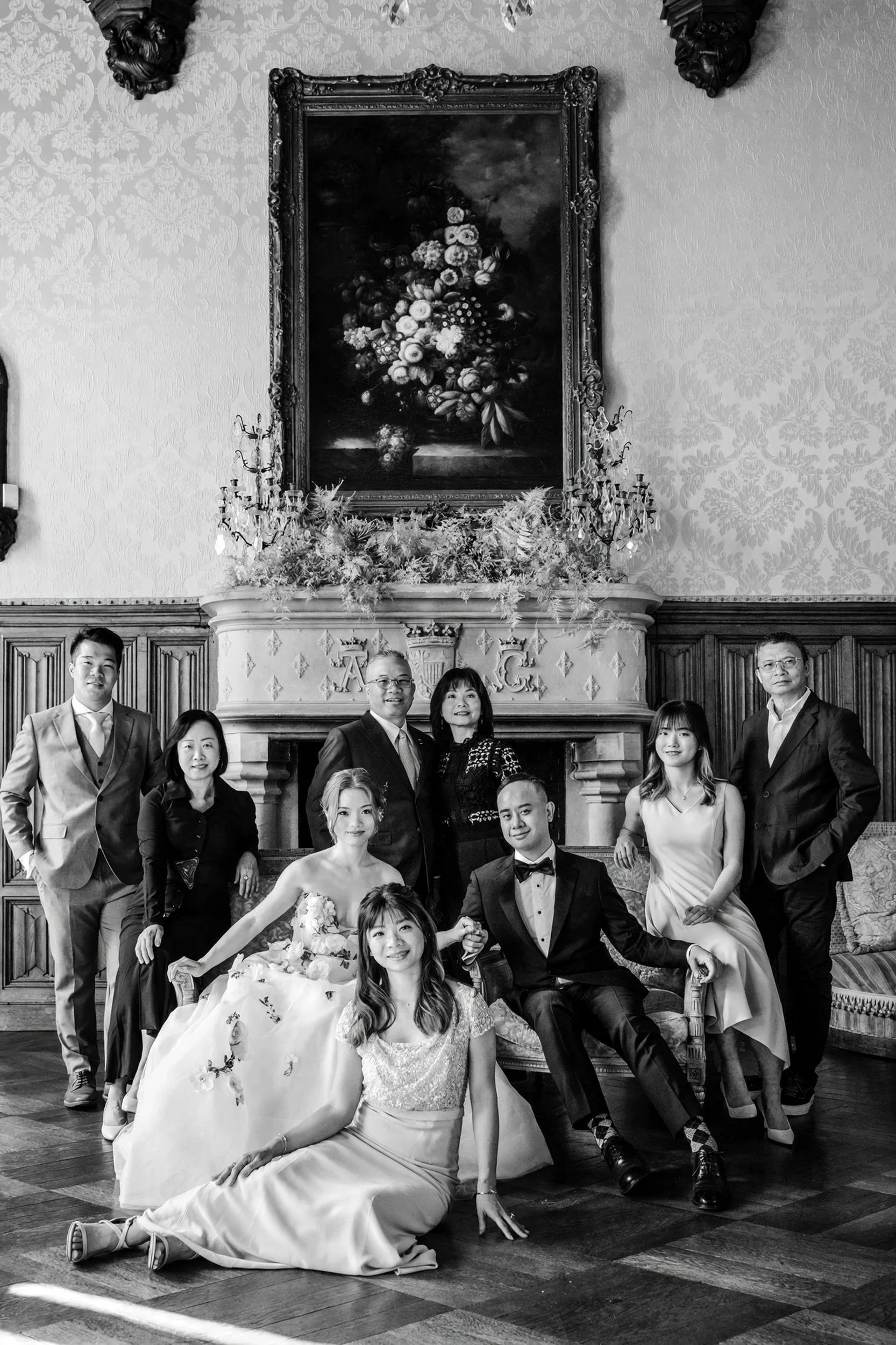 Editorial fine art black and white family portrait captured during a luxury destination wedding in a historic French chateau. The group is posed in the grand salon featuring original damask wallpaper, ornate stone fireplace with monogram, and a class