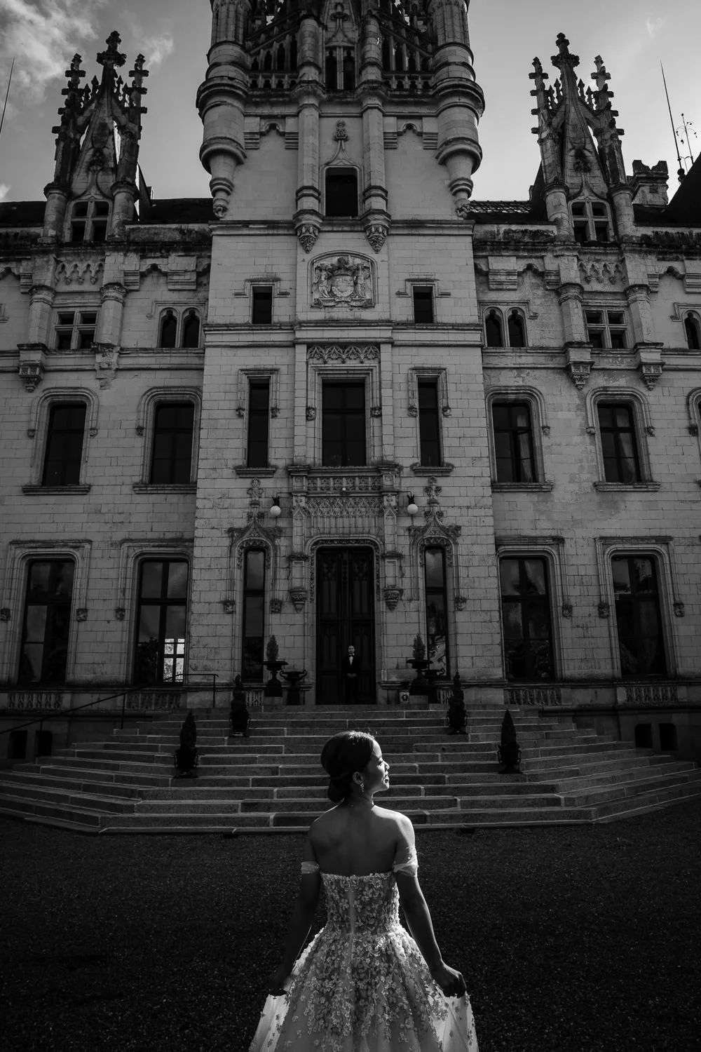 A woman in an ornate wedding dress stands outside a large, historic castle-like building with steps leading up to the entrance, and a man in formal attire standing near the door.