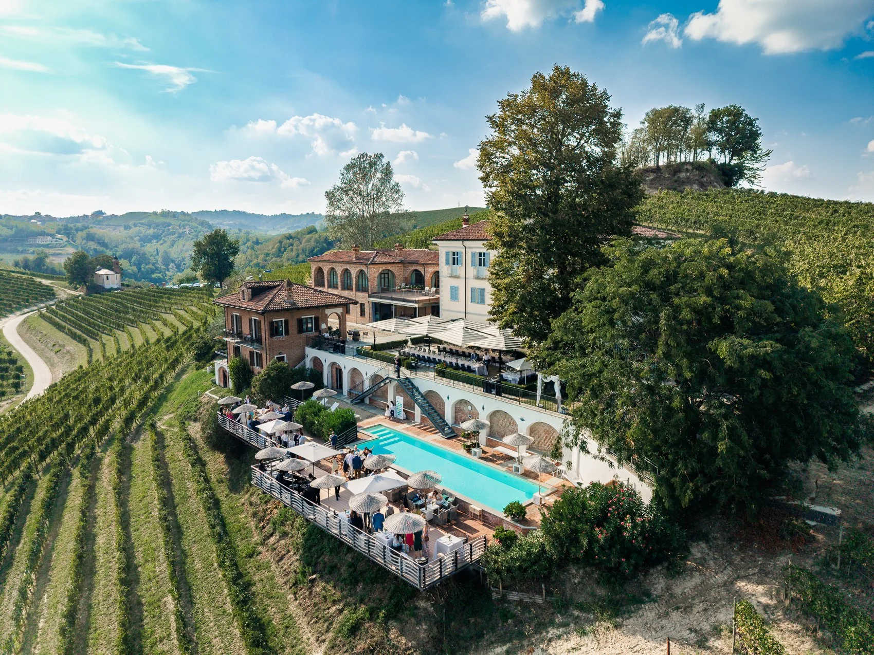 Aerial view of a scenic hillside winery estate with a swimming pool, outdoor dining area, and terraced vineyards under a blue sky with scattered clouds.