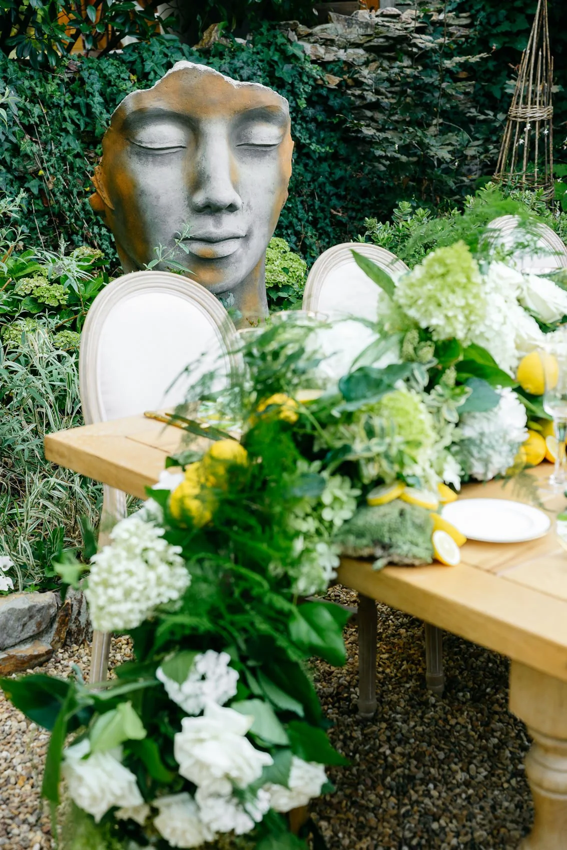 A garden scene with a large artistic face sculpture with closed eyes, surrounded by lush greenery and a wooden table decorated with white and yellow flowers and citrus slices.