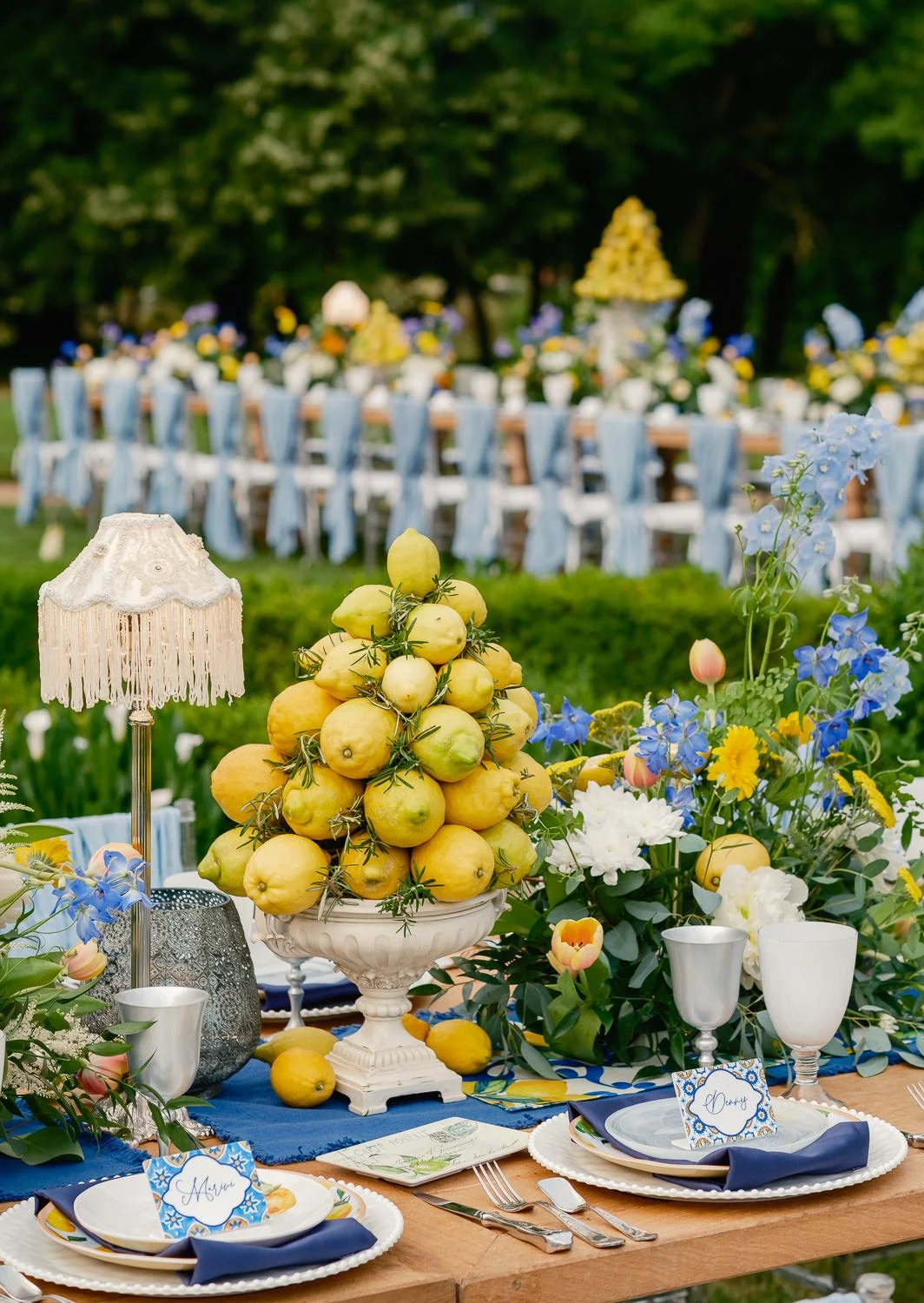 Elegant outdoor dining table decorated with lemons, flowers, and dinnerware, with a long banquet in the background for a celebration.