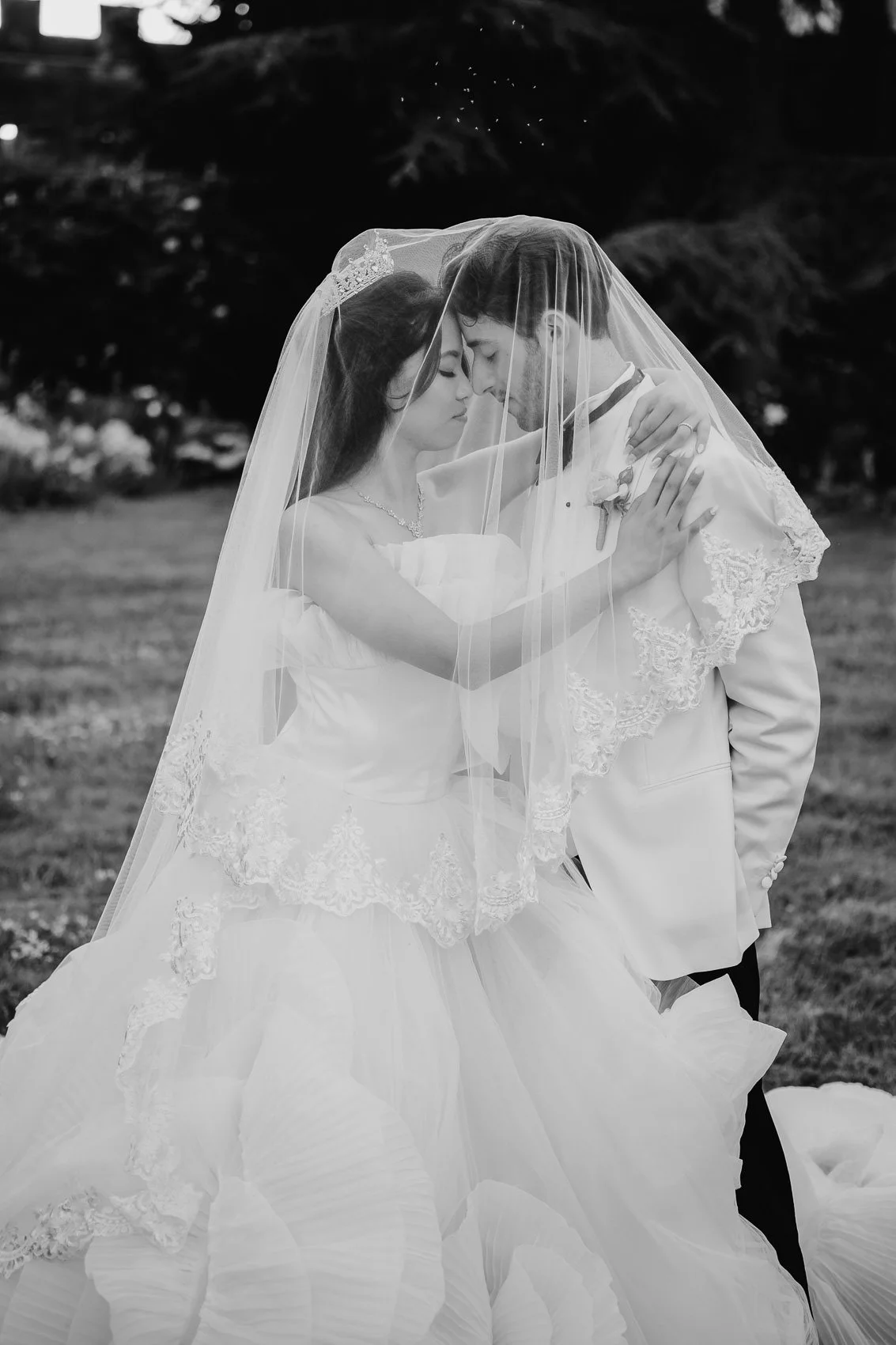 Black and white photograph of a bride and groom embracing each other with their foreheads touching, under a veil, outdoors with trees in the background.