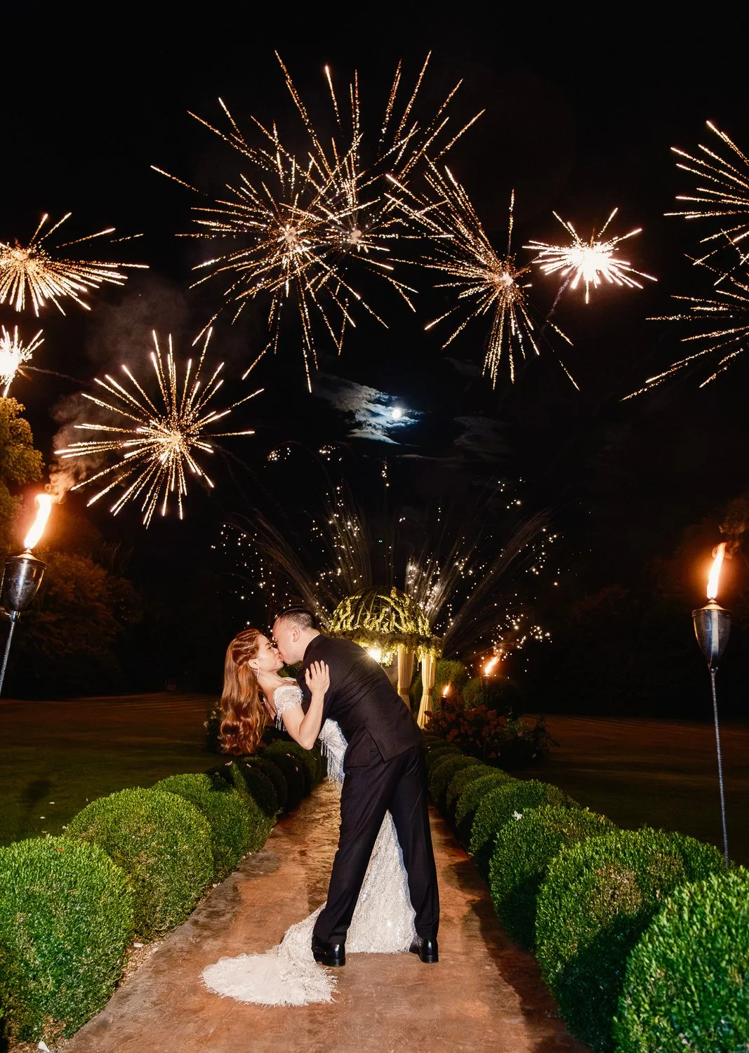 A newlywed couple kissing on a path surrounded by trimmed bushes during a fireworks display at night, with a full moon in the sky.