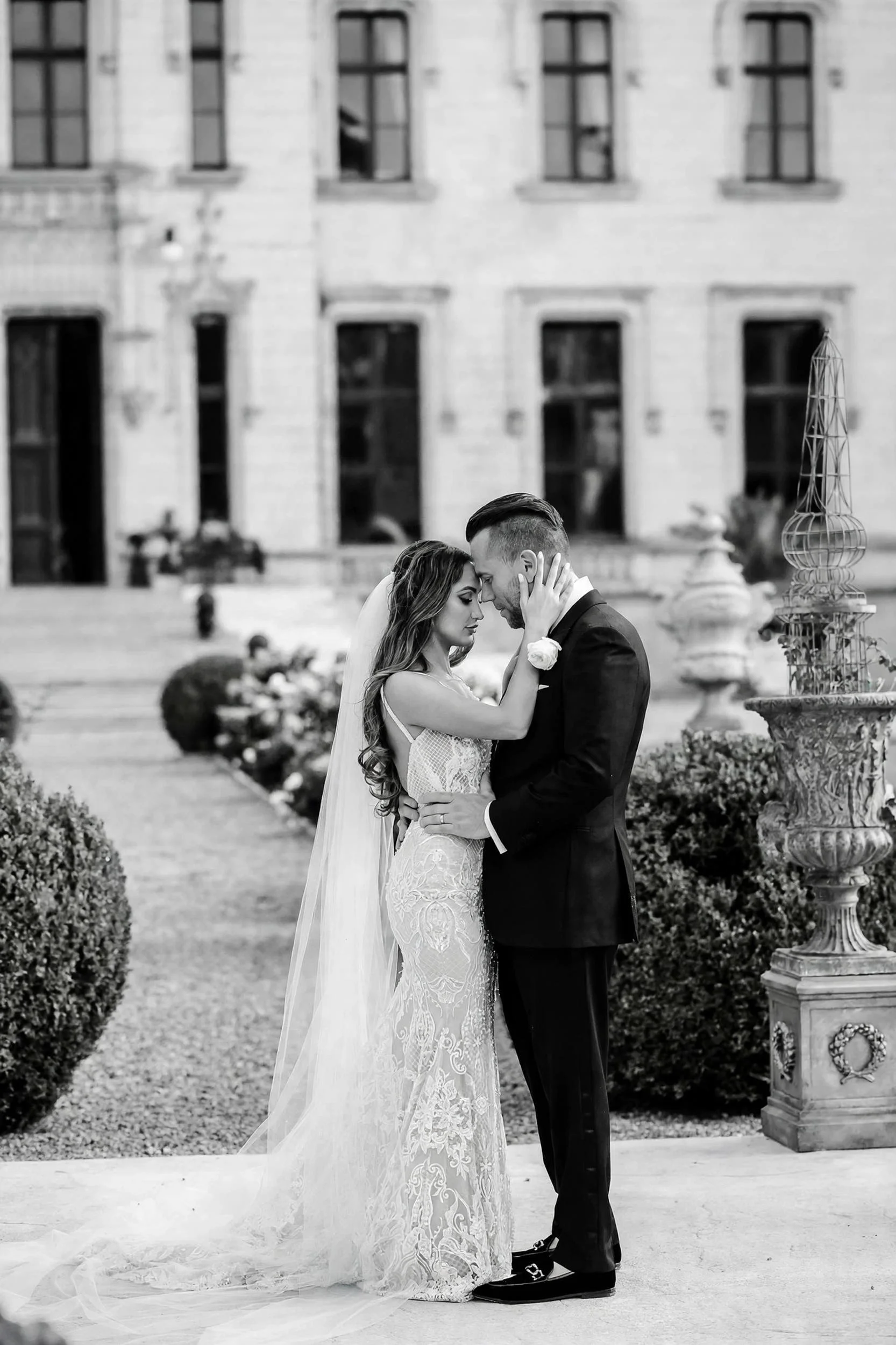 Fine art black and white wedding portrait of a bride and groom sharing an intimate moment in the gardens of a luxury French chateau, bride in lace mermaid dress with long veil