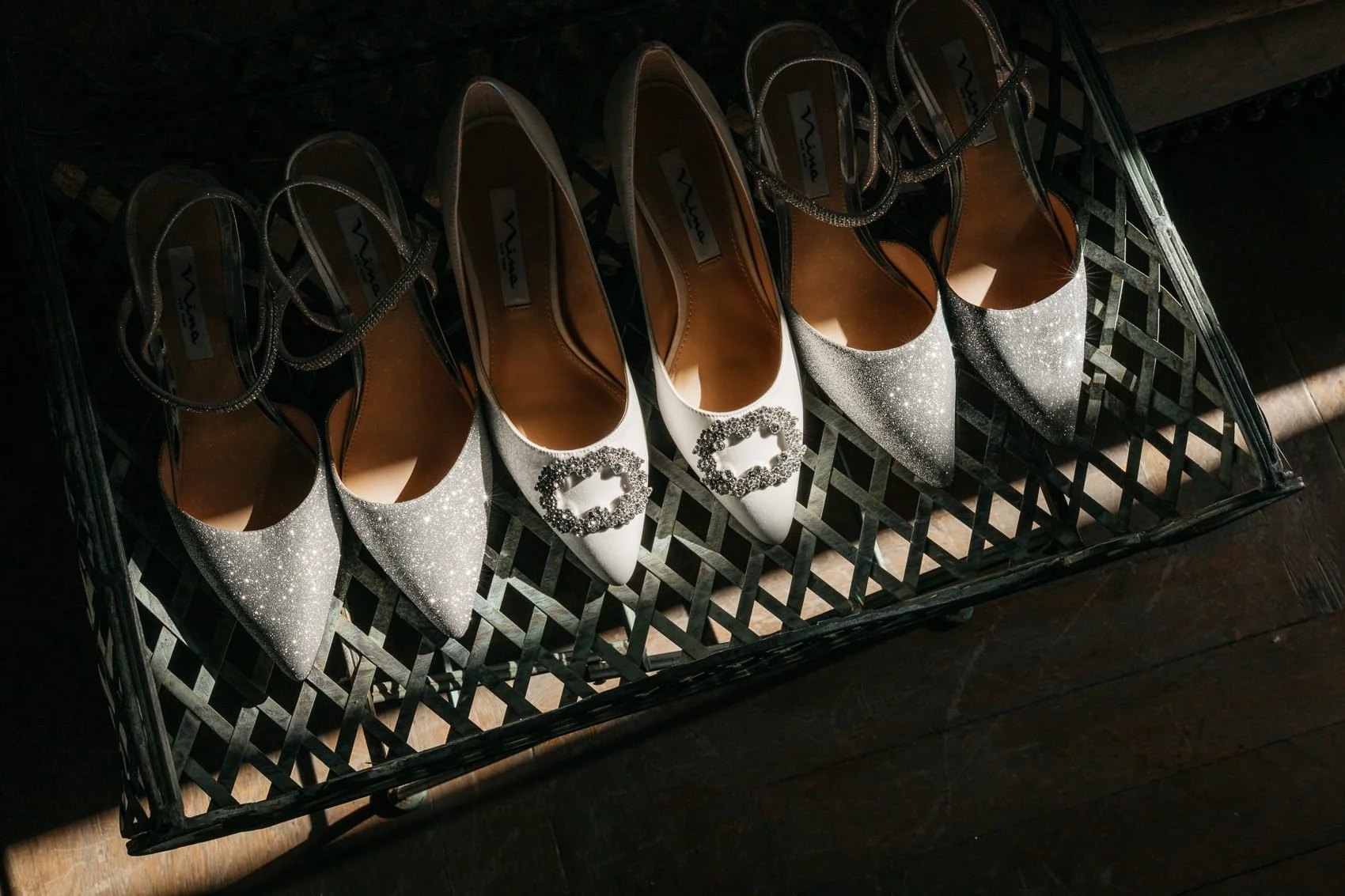 Four pairs of high-heeled shoes on a black wire basket, with two pairs having glittery silver finish and decorative buckles on the toe, and the other two pairs in shiny black finish with straps.