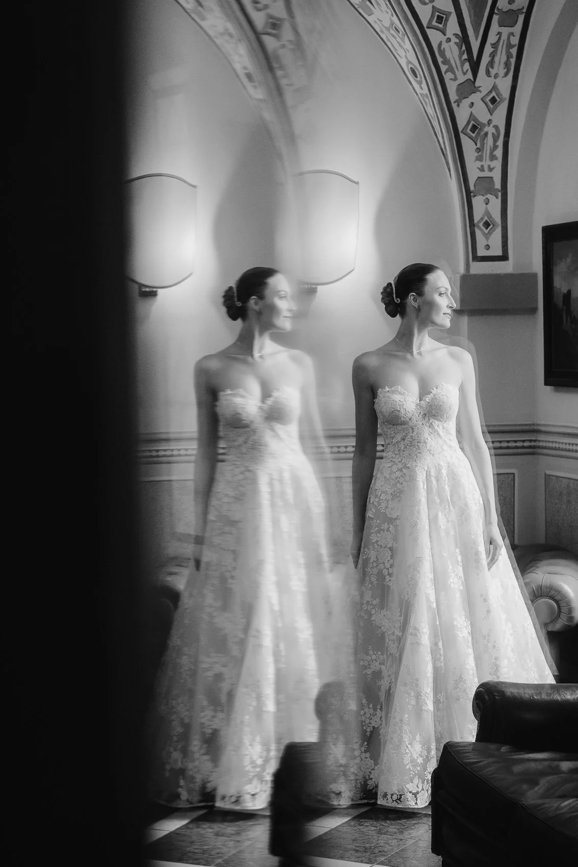 A woman in a wedding dress is standing indoors in front of a mirror, with her reflection visible. The room has ornate decorative details and two wall-mounted lights. She has her hair styled in an elegant bun and is smiling slightly, looking to the si