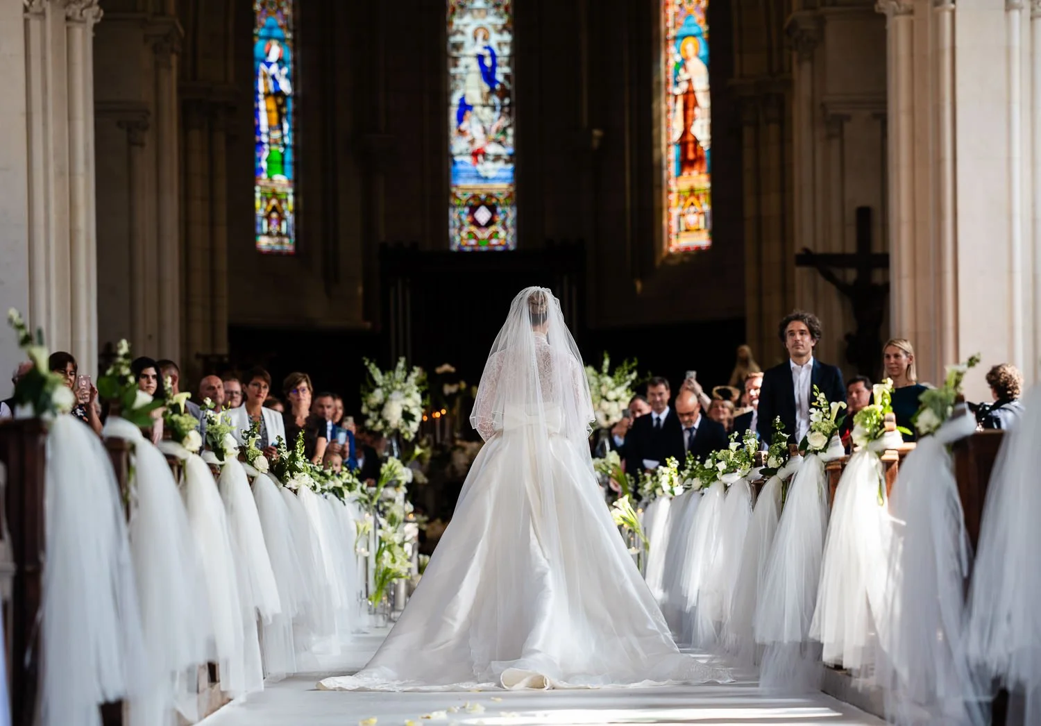 A bride with a veil and wedding dress standing at the altar during a wedding ceremony inside a church with stained glass windows and floral decorations, surrounded by guests seated in pews.