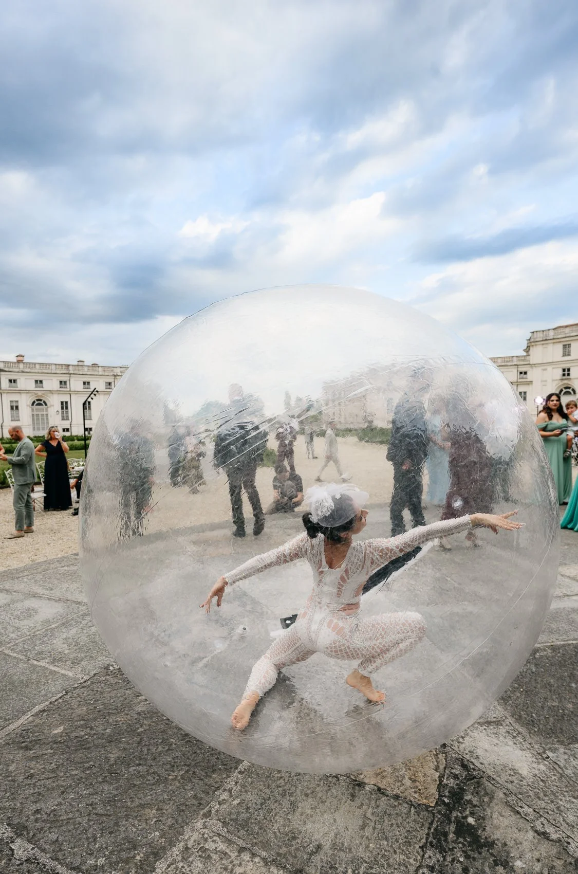 A woman dressed in a white lace outfit and white headpiece is inside a large clear plastic inflatable sphere, striking a pose with one arm extended and the other bent behind her, during an outdoor event. Several people are seen walking or standing in