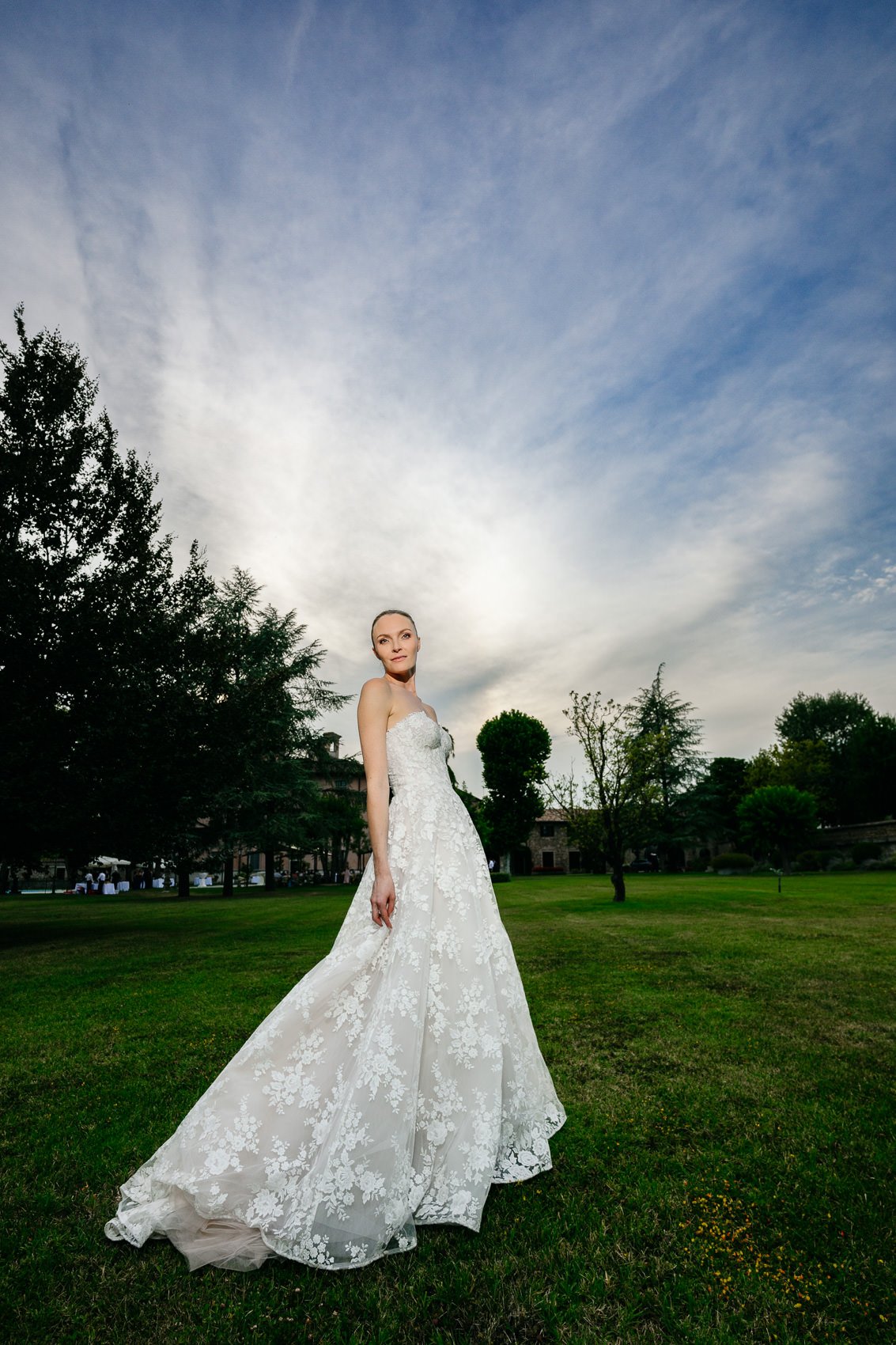 A woman in a white lace wedding dress standing on a grassy lawn with trees and buildings in the background under a partly cloudy sky.