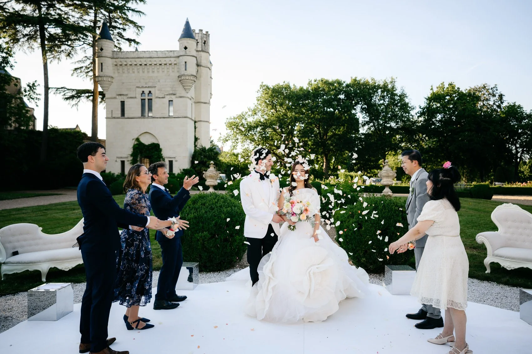 A wedding ceremony outdoors with a castle in the background. The bride and groom are standing in the center, with the bride holding a bouquet. Guests are standing on either side, celebrating as flower petals are showered around them.