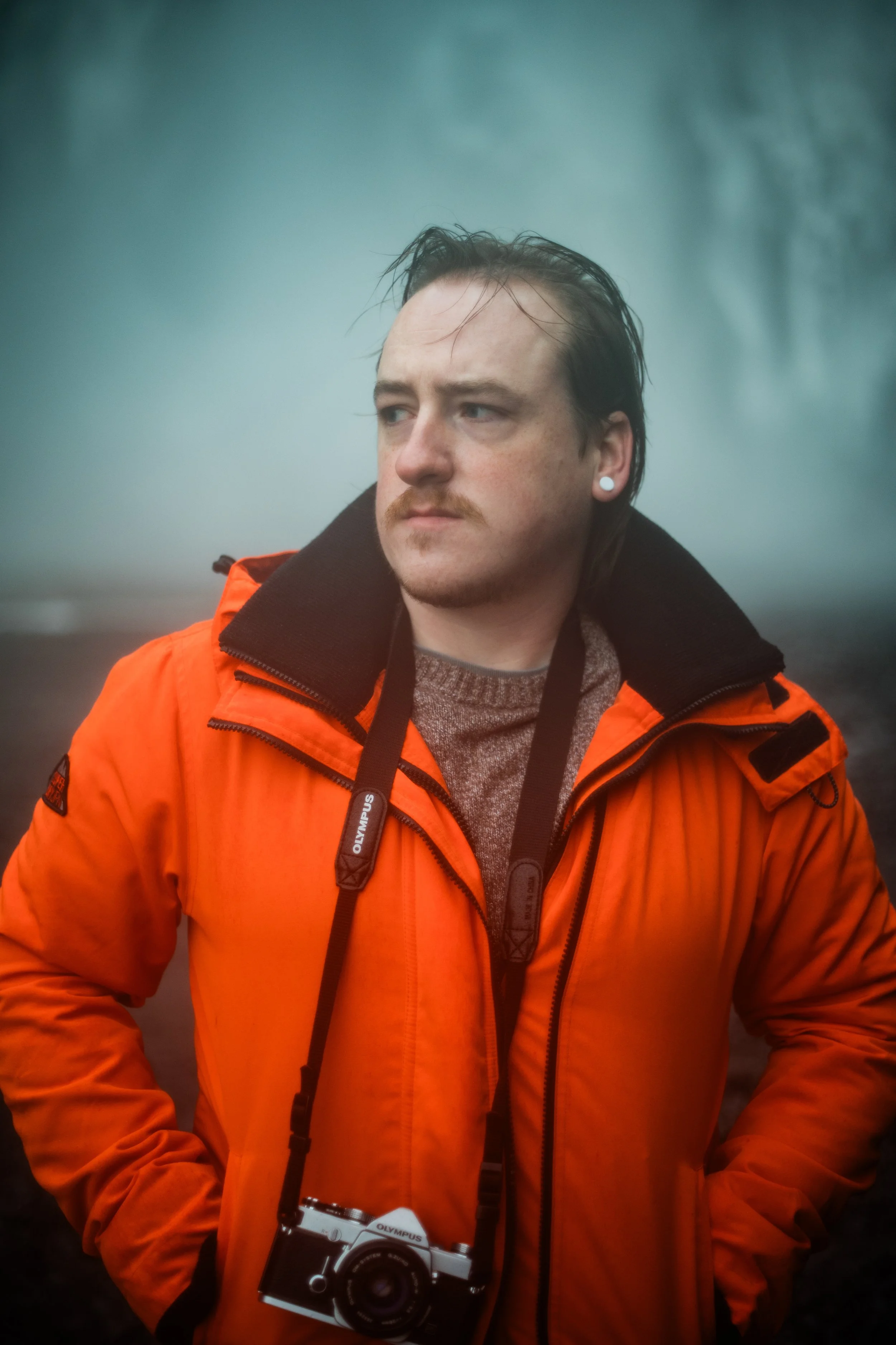 A man with dark, wet hair and a mustache wearing an orange jacket and a camera around his neck, standing outdoors in a foggy or misty environment.