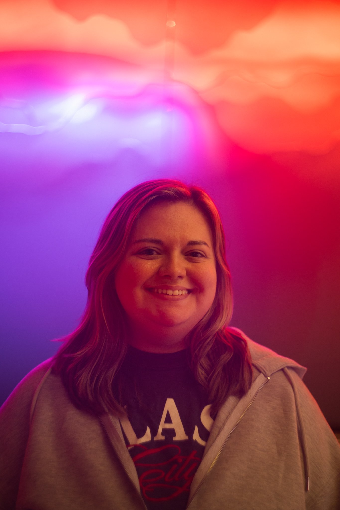 A smiling woman with shoulder-length hair in front of a colorful, neon-lit background with shades of pink, purple, and orange.