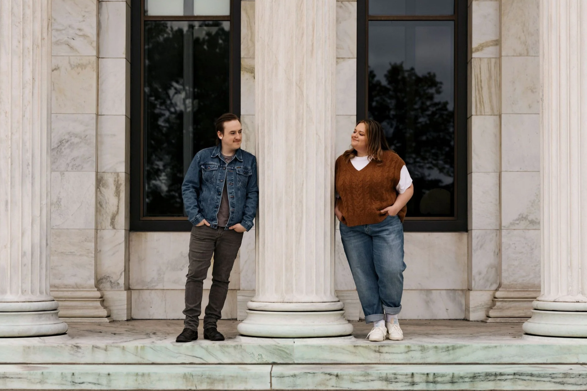 A man and woman standing on a marble steps in front of a building with large windows and white columns, engaged in conversation.