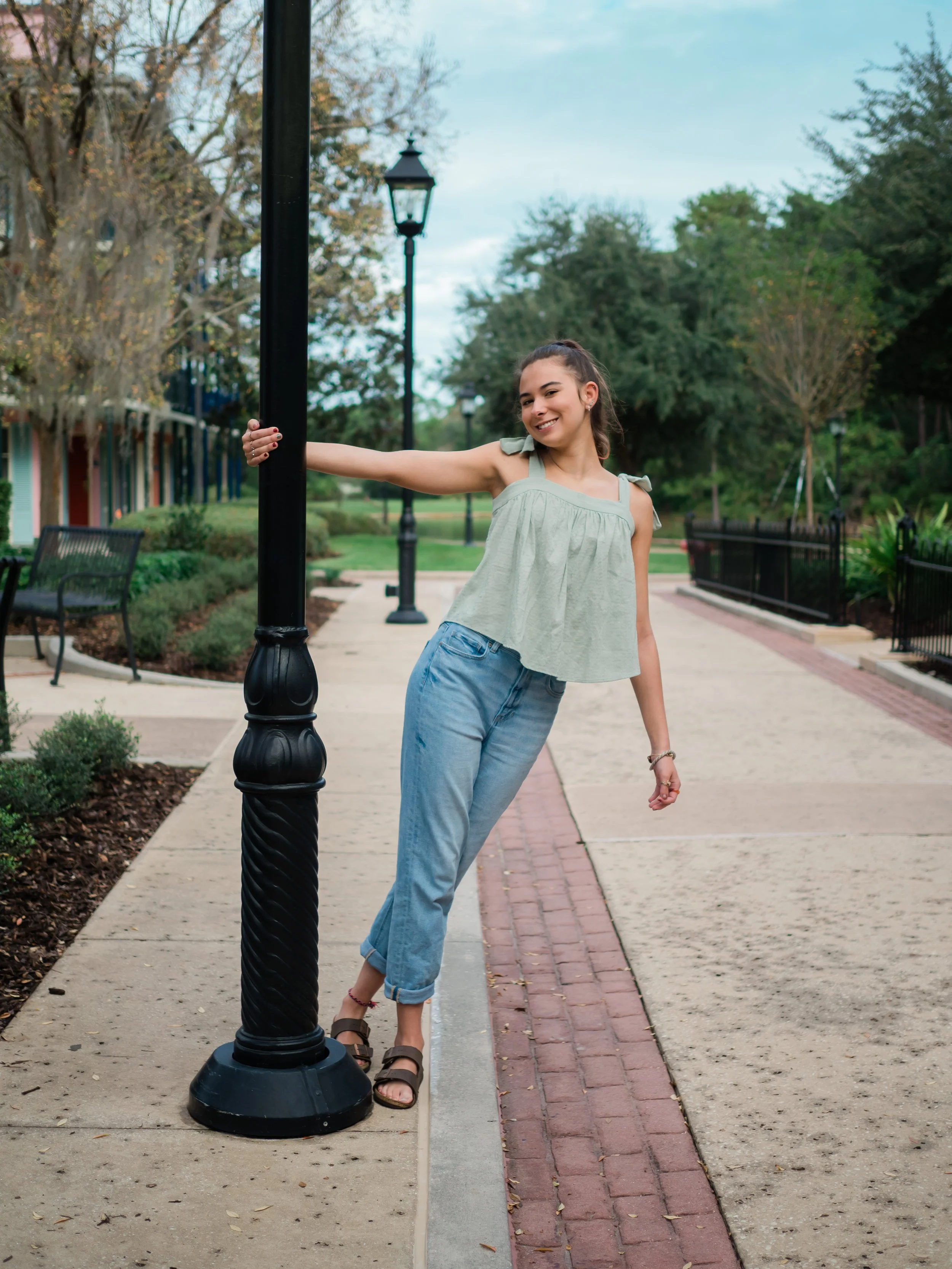 A young woman leaning on a black lamp post on a paved sidewalk in a park-like setting, smiling and looking to the side. She is wearing a light green sleeveless top, light blue rolled-up jeans, and sandals, with trees and lamps in the background.