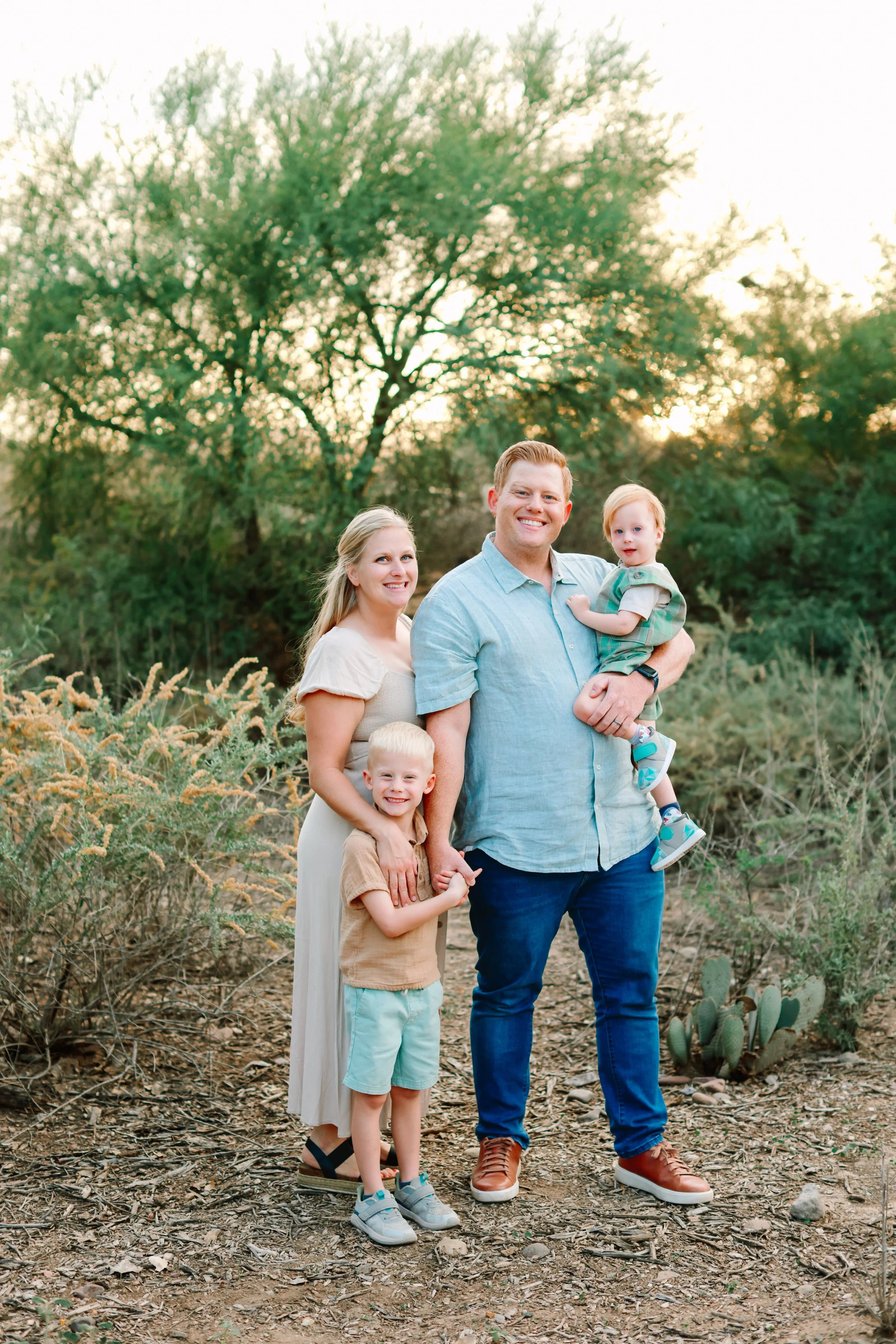 A happy family of four standing outdoors on a dirt path surrounded by desert vegetation, including cacti and shrubs, with trees in the background and the sun setting behind them.