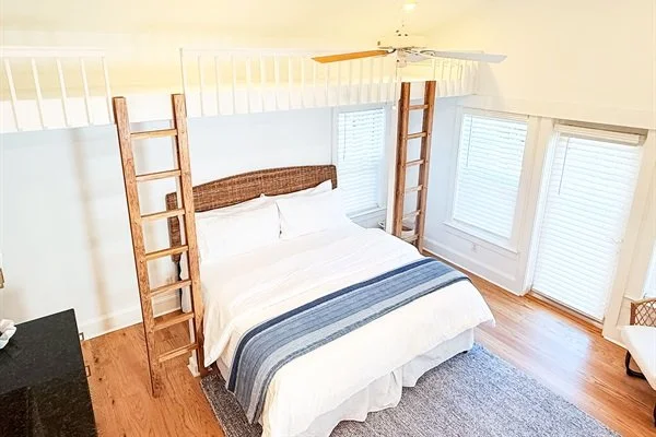 Bedroom with a white bed, wooden loft ladder, ceiling fan, hardwood floors, and large windows with blinds.