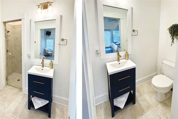 A bathroom with a navy blue vanity with a white sink, gold faucet, and a mirror above it. The bathroom has light-colored tiled floors and walls, with a shower area visible through an open door. There is a toilet next to the vanity on the right.