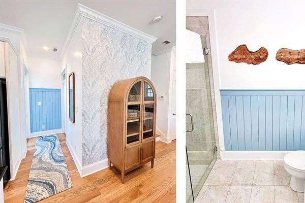 Interior view of a home with hardwood floors, blue wainscoting walls, a patterned rug, a wooden cabinet, and a bathroom with tile flooring, a glass shower, and decorative wooden wall hangings.