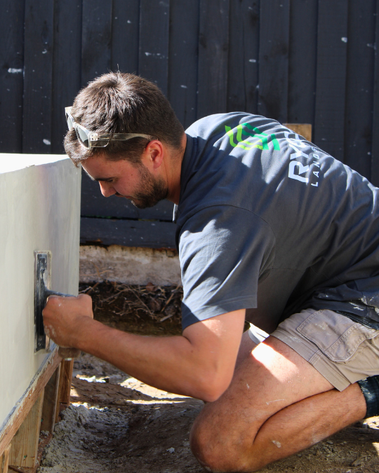 Man working on the exterior of a house, kneeling on the ground near a wooden fence, wearing sunglasses and a gray T-shirt.