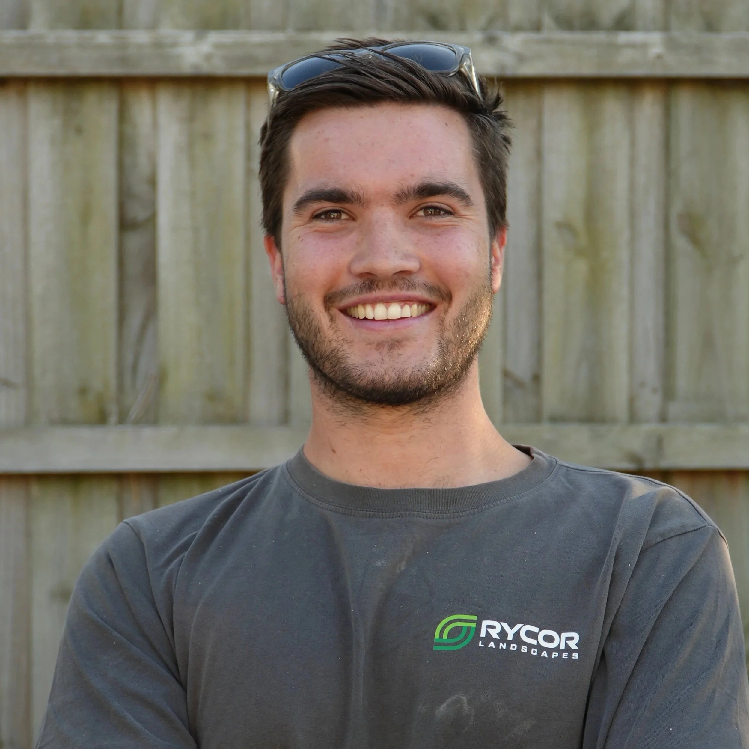 Close-up of a man smiling, wearing sunglasses on his head and a dark gray T-shirt with a logo reading 'RYCOR Landscapes' on the front, standing in front of a wooden fence.