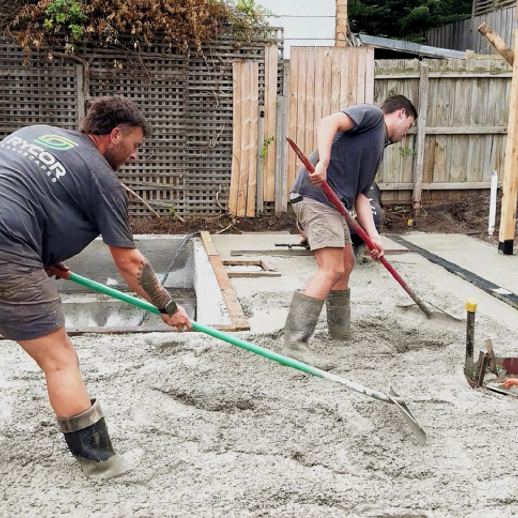Two men working on a construction site pouring concrete, with a wooden fence and trees in the background.