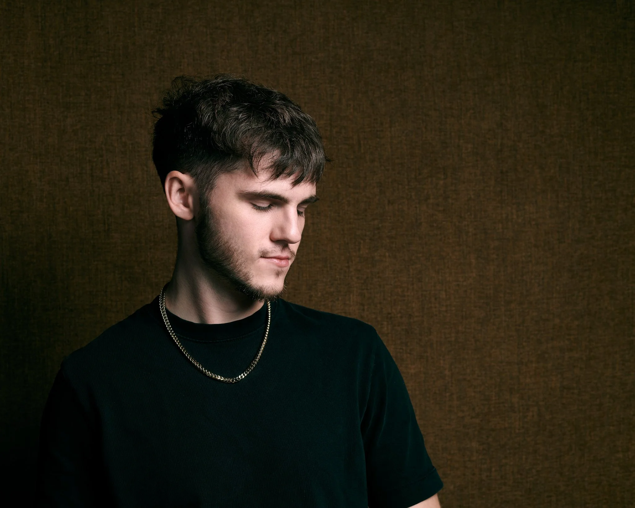 Portrait studio d'un jeune homme en t-shirt noir et chaîne dorée, regard baissé devant un fond texturé brun, photographié par Montreal Headshot à Montréal
