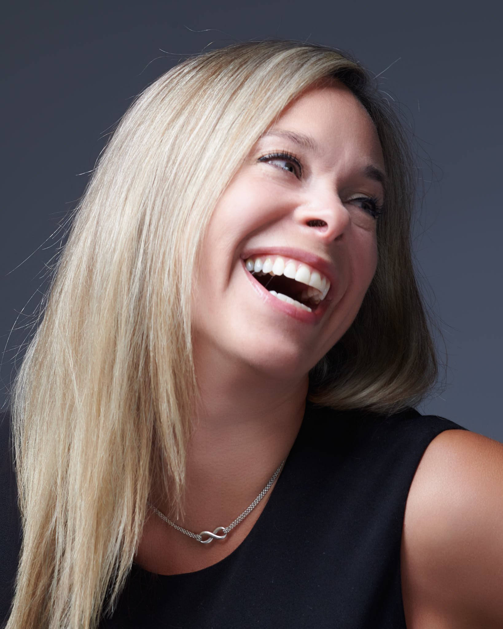 Femme souriante avec des cheveux blonds et une peau claire, portant un t-shirt noir, un collier en chaîne et un fond gris.