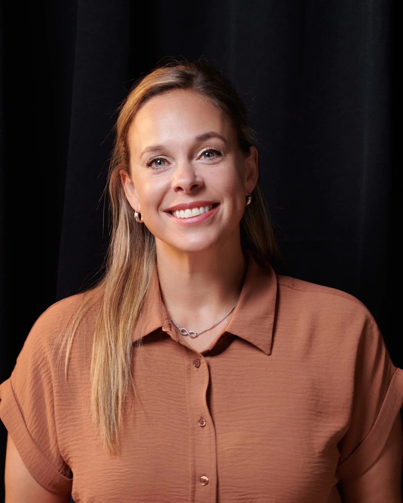 Une femme souriante portant une chemise marron clair avec un collier en forme de symbole infini, contre un fond noir.
