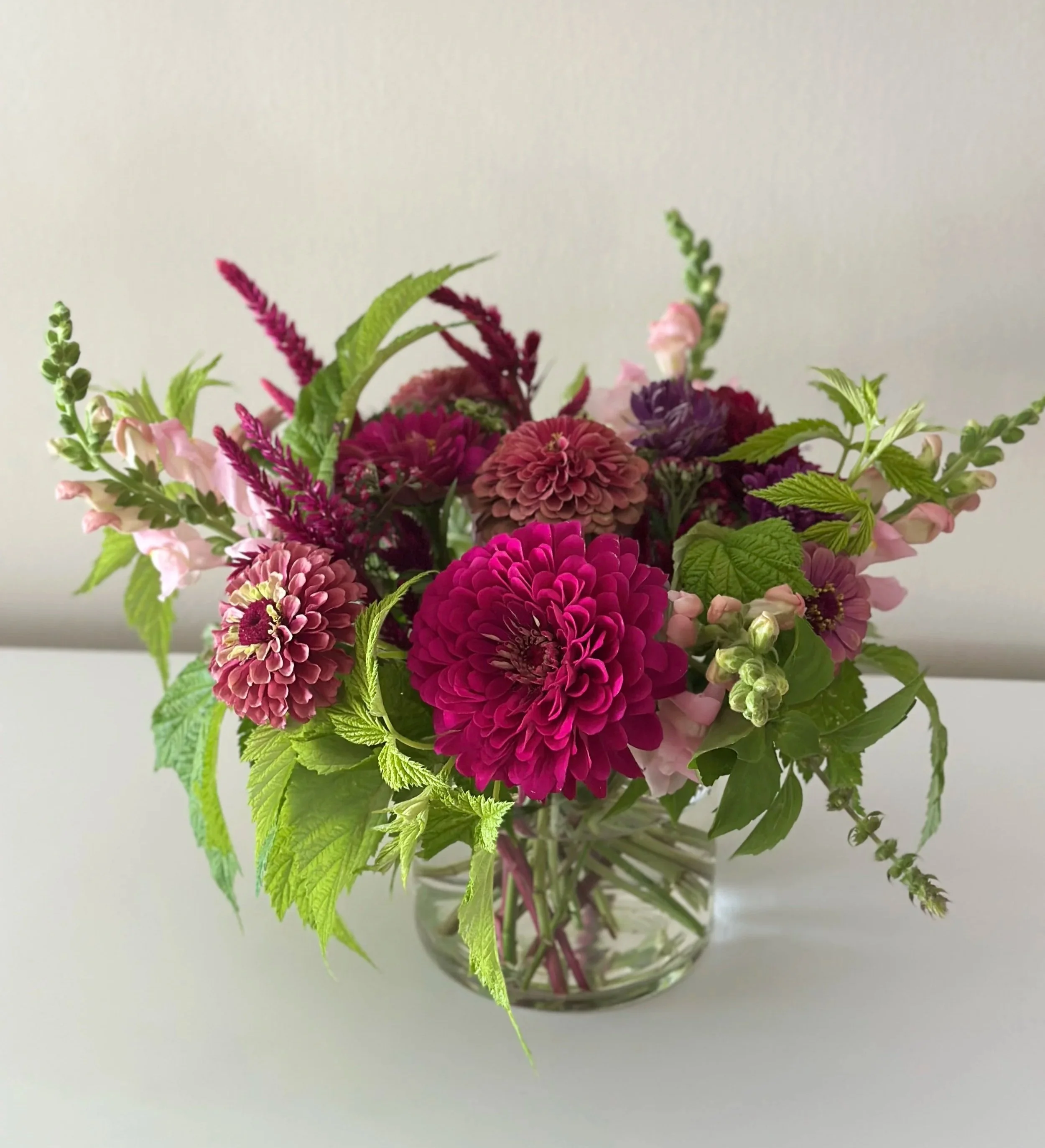 Centerpiece flower arrangement in a glass vase with dark wine, purple, and green flowers and leaves on a white background.