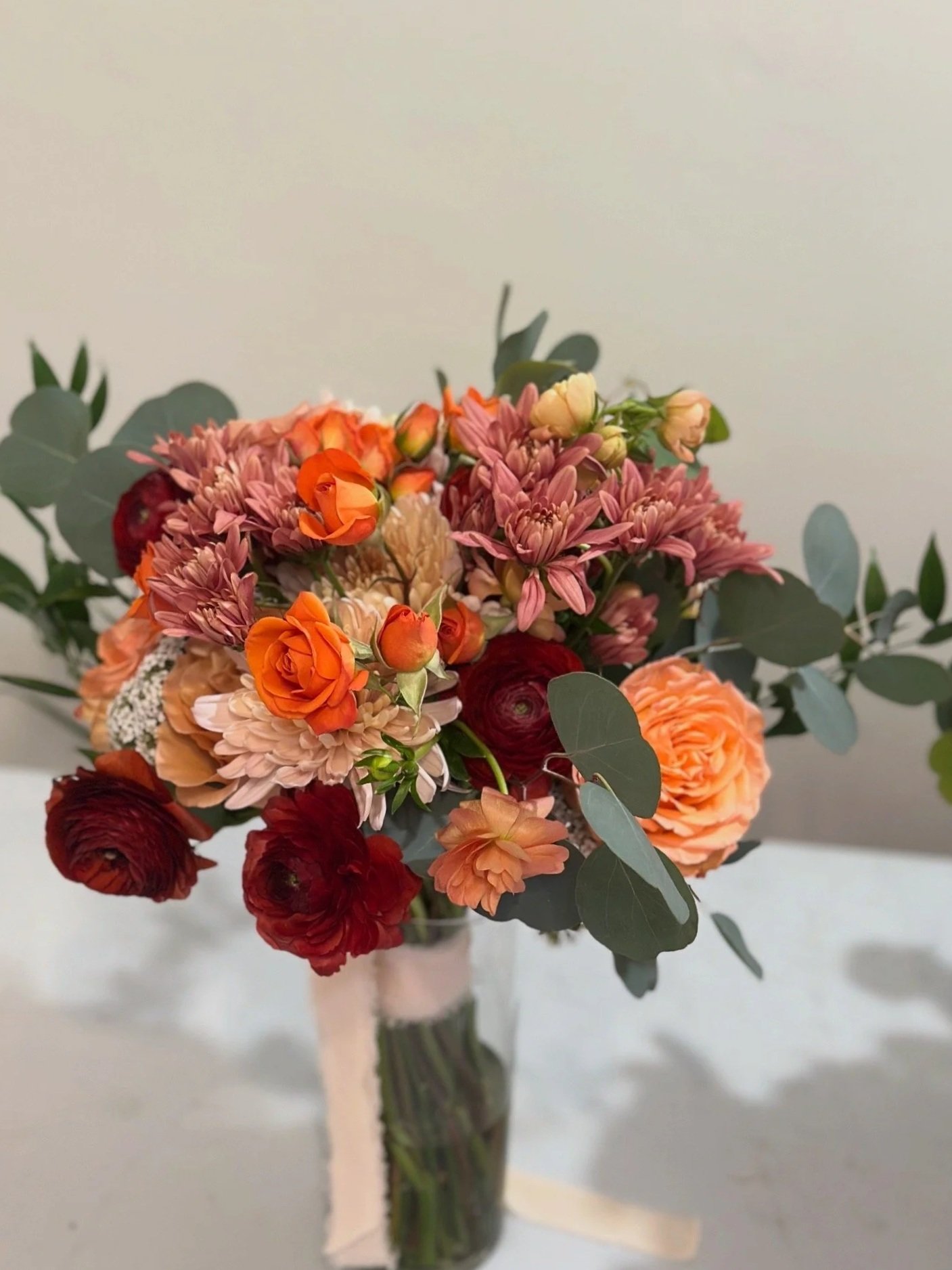 A bridal bouquet of orange, terracotta, burgundy, peach, and cream  flowers in a tall glass vase with eucalyptus leaves and a silk chiffon ribbon.