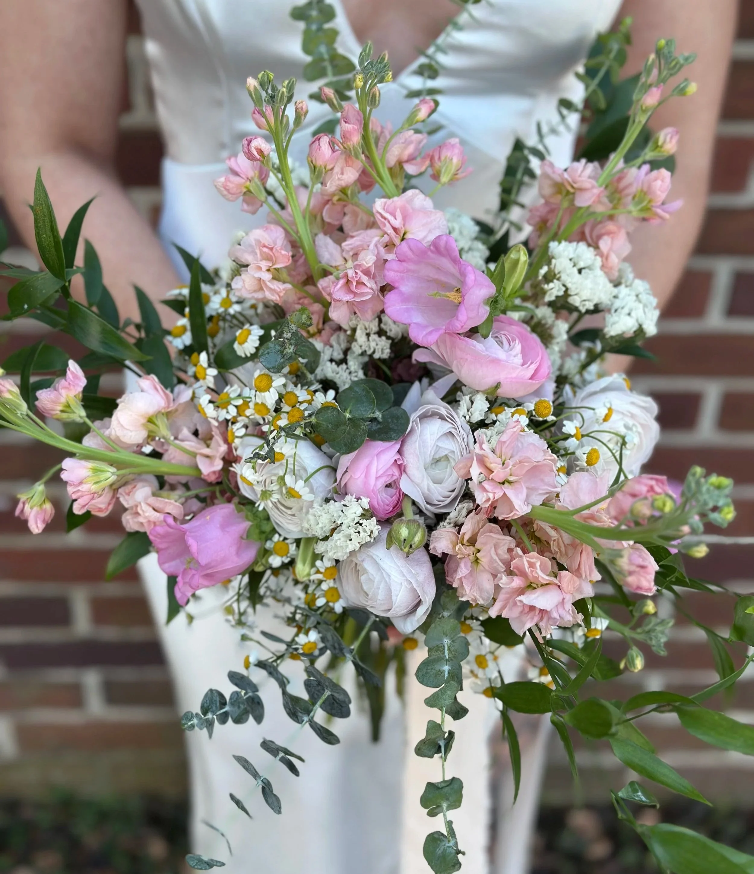 Close-up of a bouquet o a bride holding a pink, white, and lavender wedding bouquet with green leaves, against a brick background.