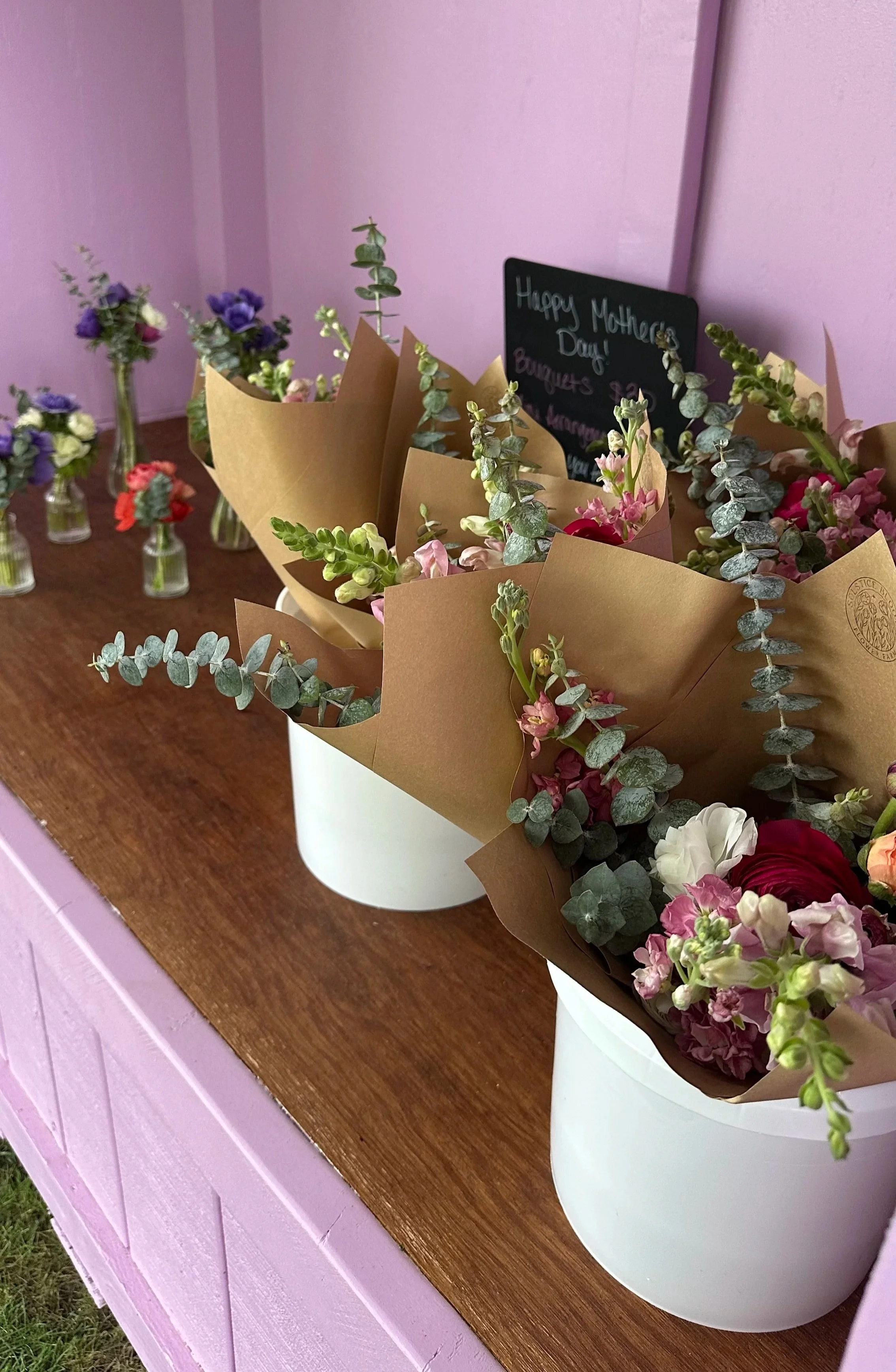 Bouquets of colorful flowers wrapped in brown paper inside white buckets, placed on a wooden surface against a pink wall of a farmstand, with a chalkboard thanking customers.