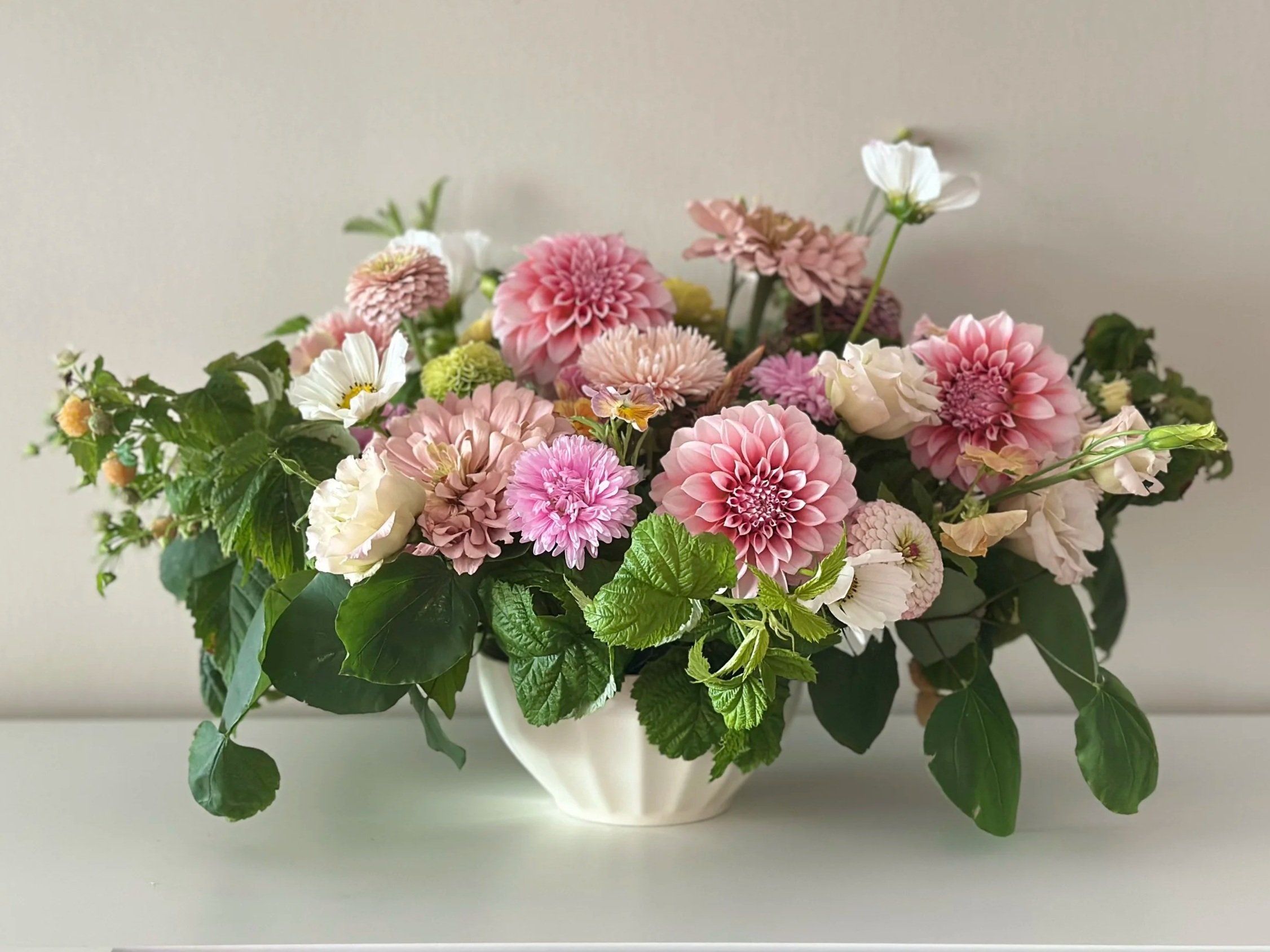 A table arrangement of pink, white, and cream flowers with green leaves in a white vase, placed in a cream compote on a white surface against a plain light background.
