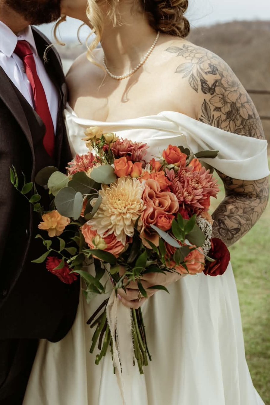 A bride holding a bouquet of orange, peach, and cream flowers with greenery, standing next to a groom in a black suit with a red tie, outdoors.