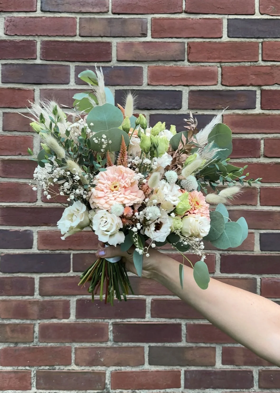 Hand holding a pastel bouquet of zinnias, dahlias, lisianthus, eucalyptus, baby's breath, and dried grasses against a brick wall