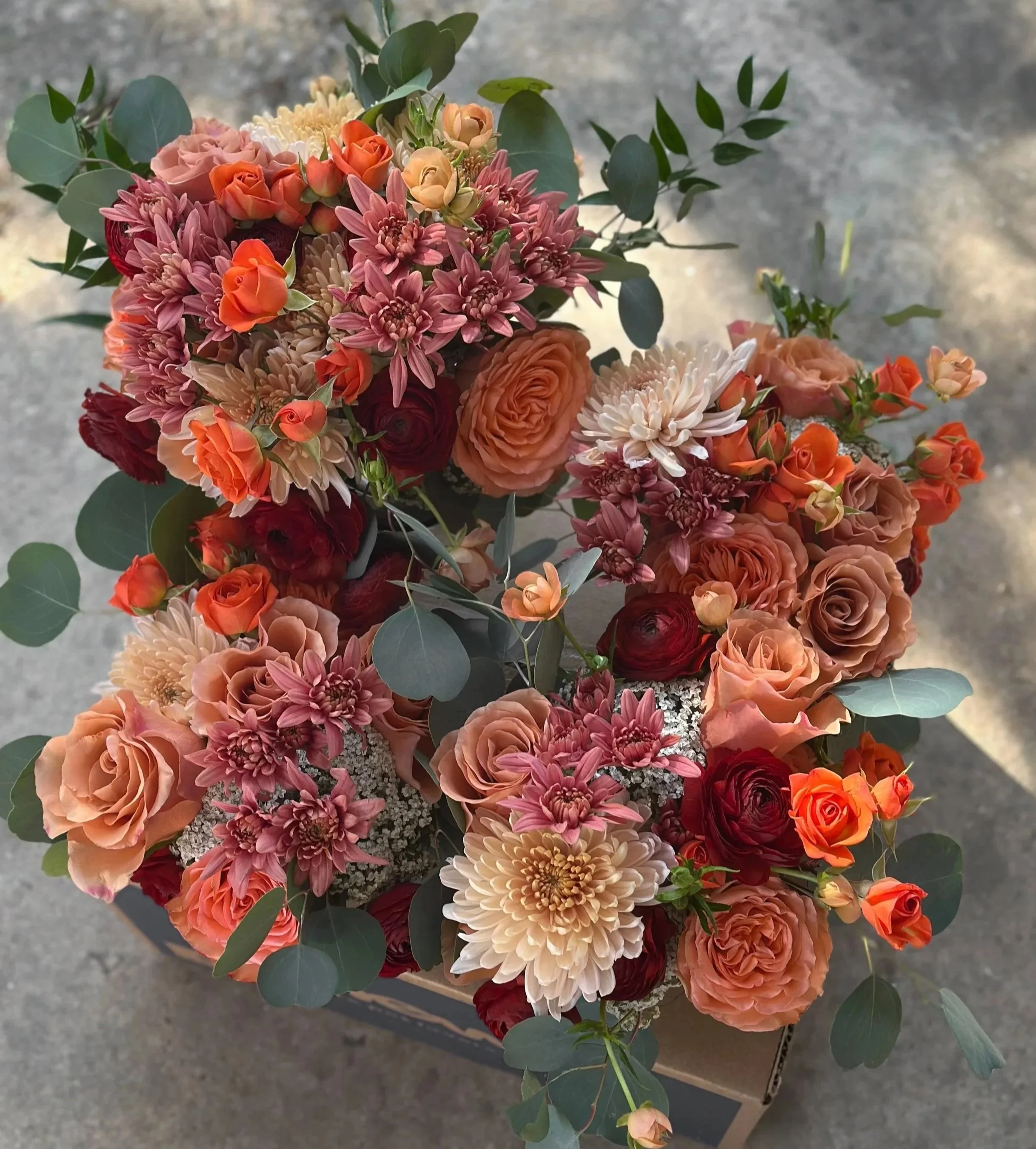 A group of wedding bridal bouquets of various peach, pink, and deep red burgundy flowers with green eucalyptus leaves packed in a box for pick up A la Carte flowers.