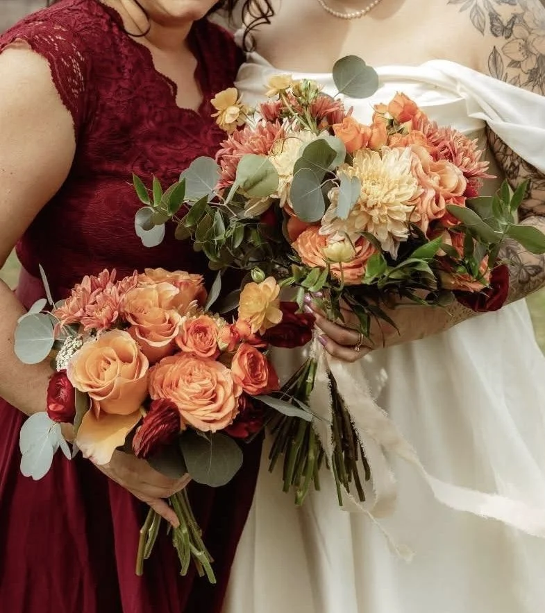 Two women holding large bouquets of peach, orange, and red flowers, including roses and dahlias, with green foliage, at a wedding or special event.